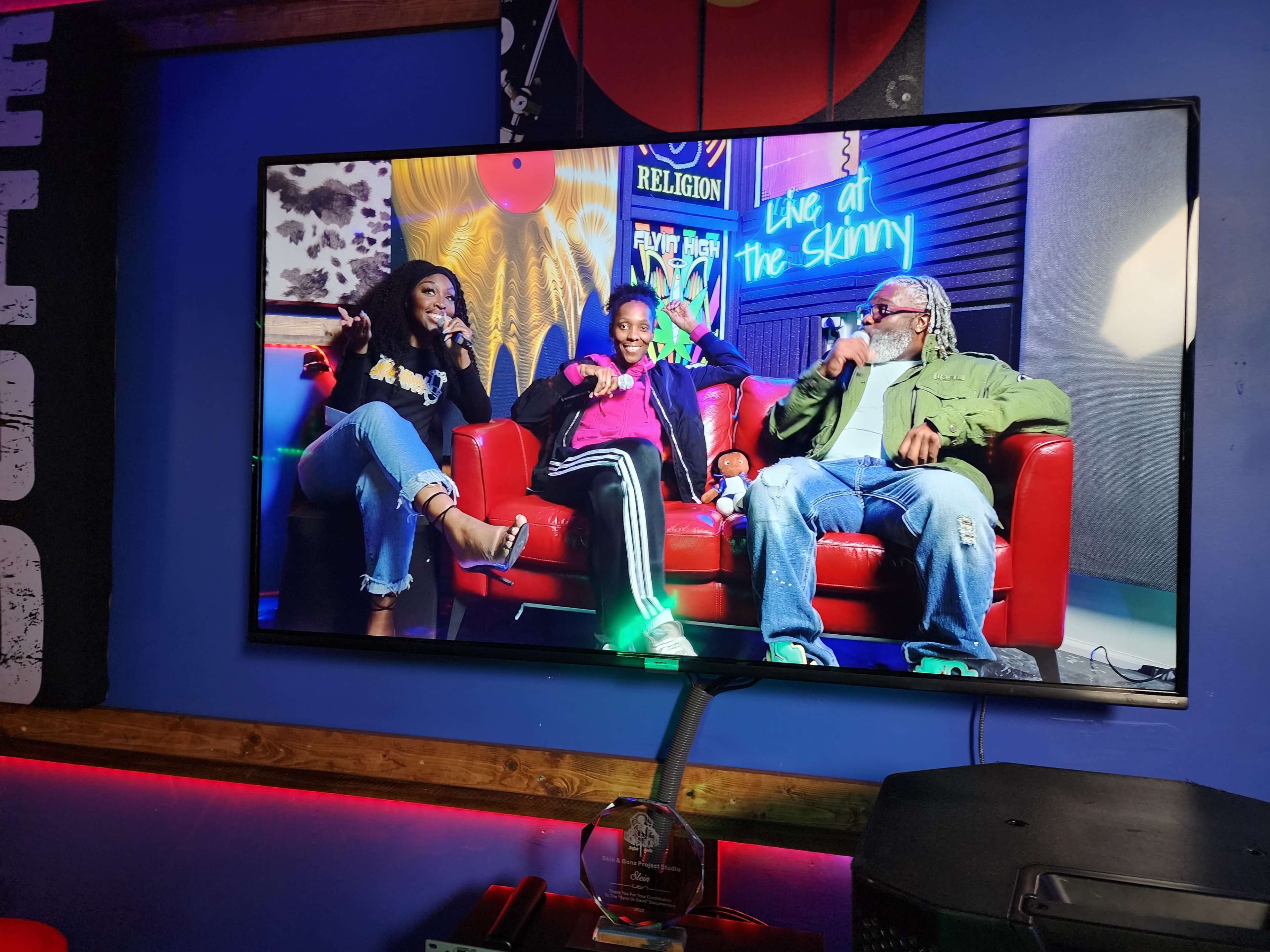 A television screen displays a live interview with three people sitting on a red couch in a colorful studio setting.
