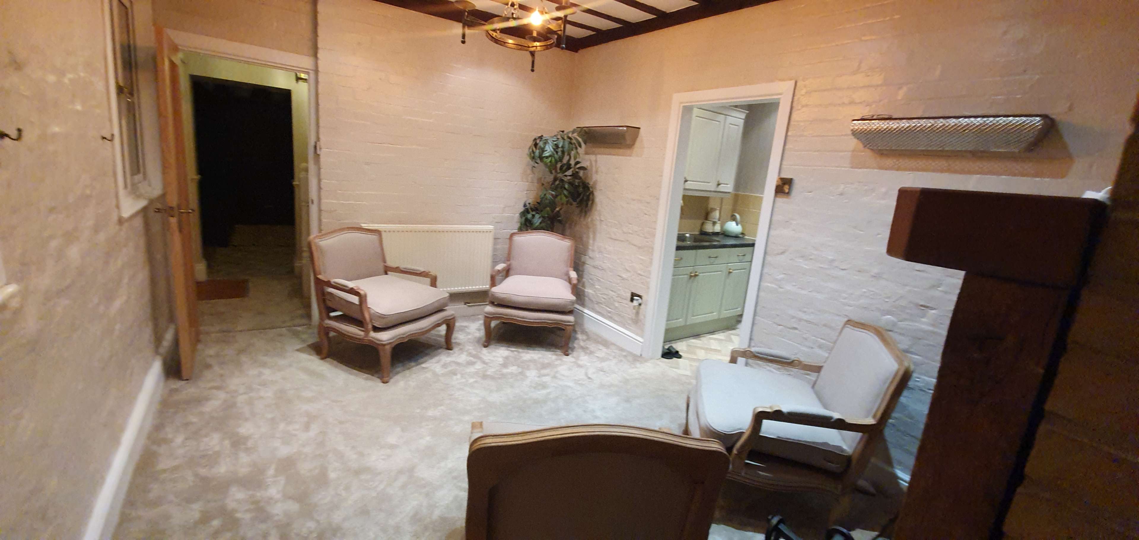A sparsely furnished room with four upholstered chairs arranged around a small, empty space and a doorway leading to a kitchen.