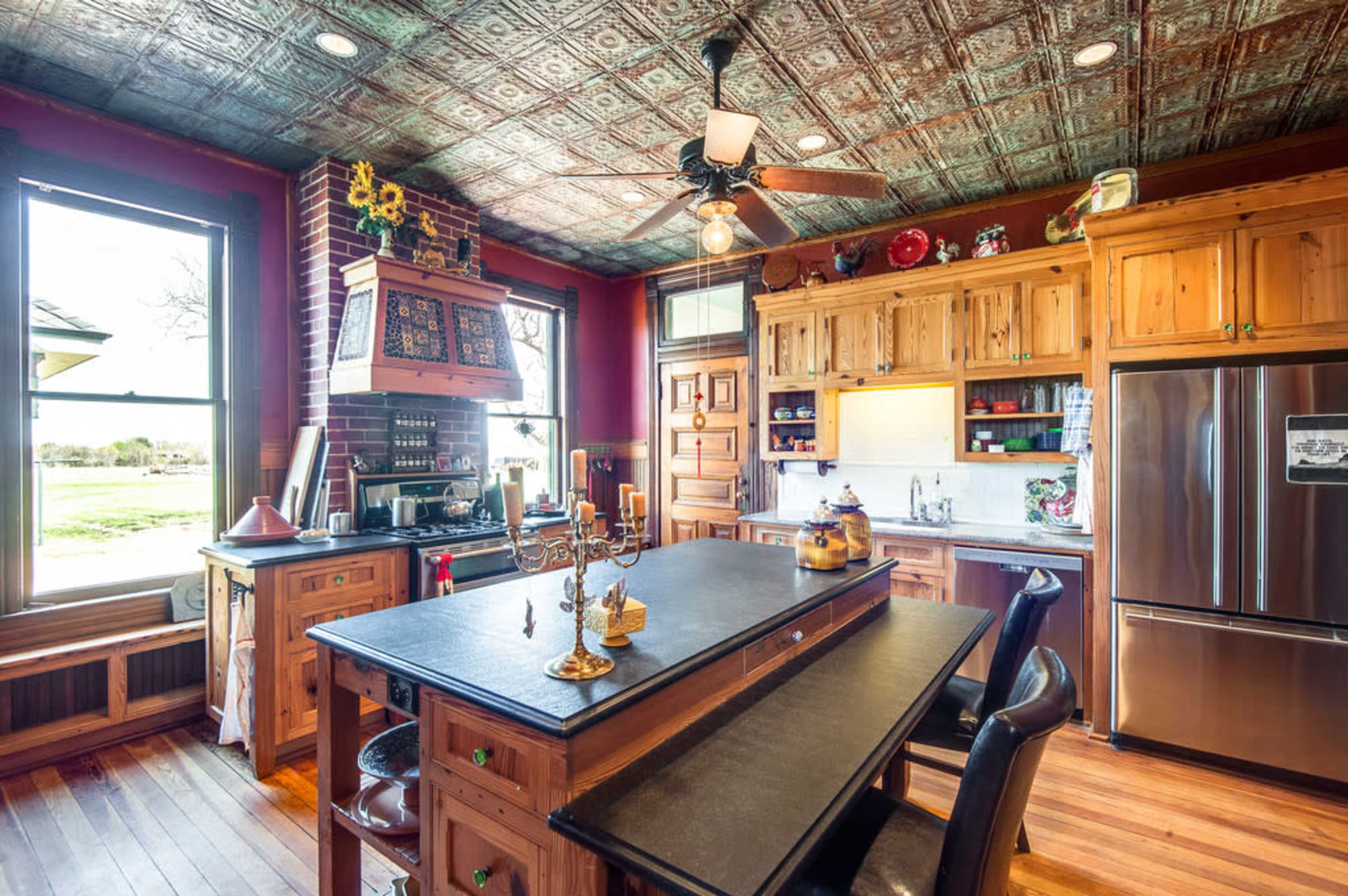 The kitchen features wooden cabinetry, a large central island, and vintage ceiling tiles, with natural light pouring in from the windows.