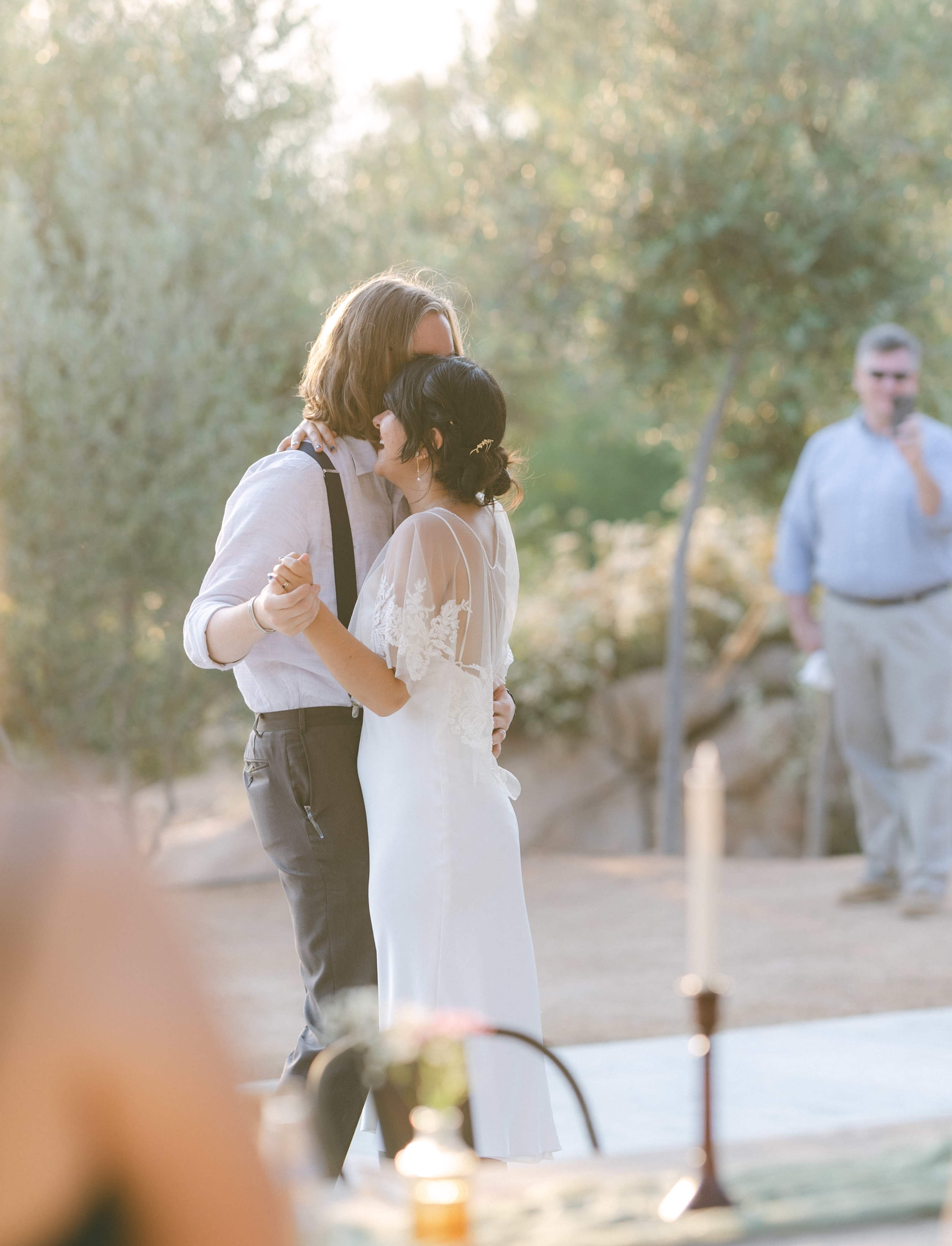 A couple dances closely together at an outdoor wedding ceremony while a guest stands in the background.
