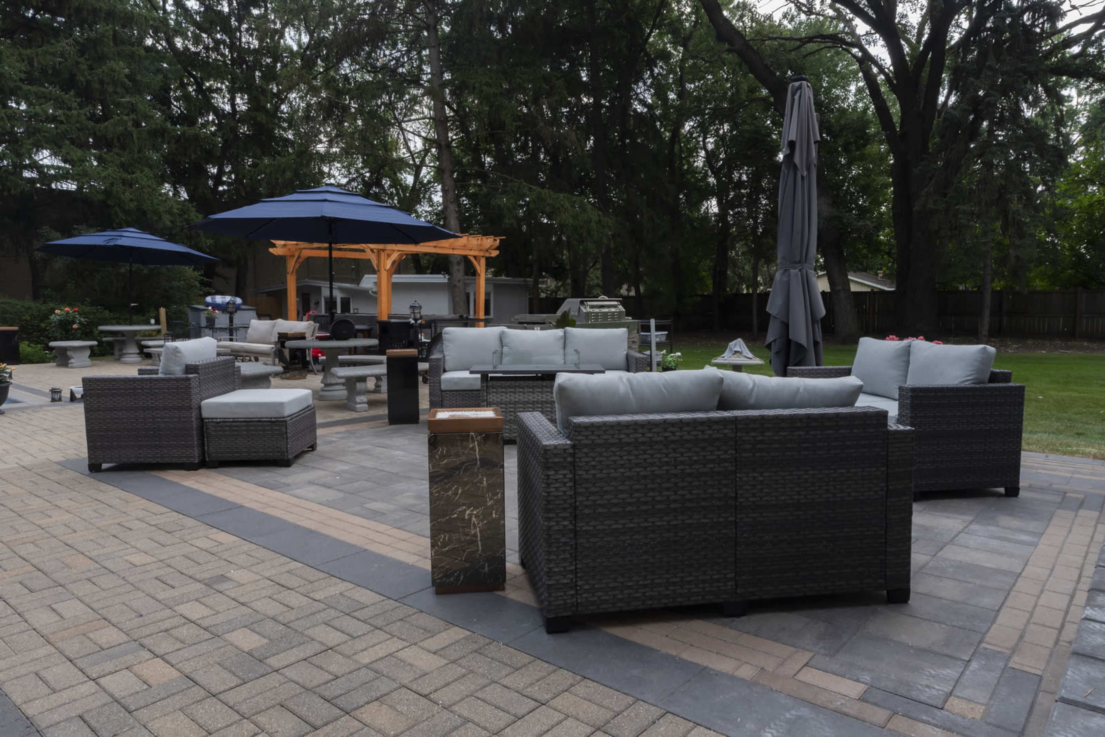 The image shows a landscaped patio area with several dark wicker seating arrangements, a wooden gazebo, umbrellas, and a fire pit on a paved surface.