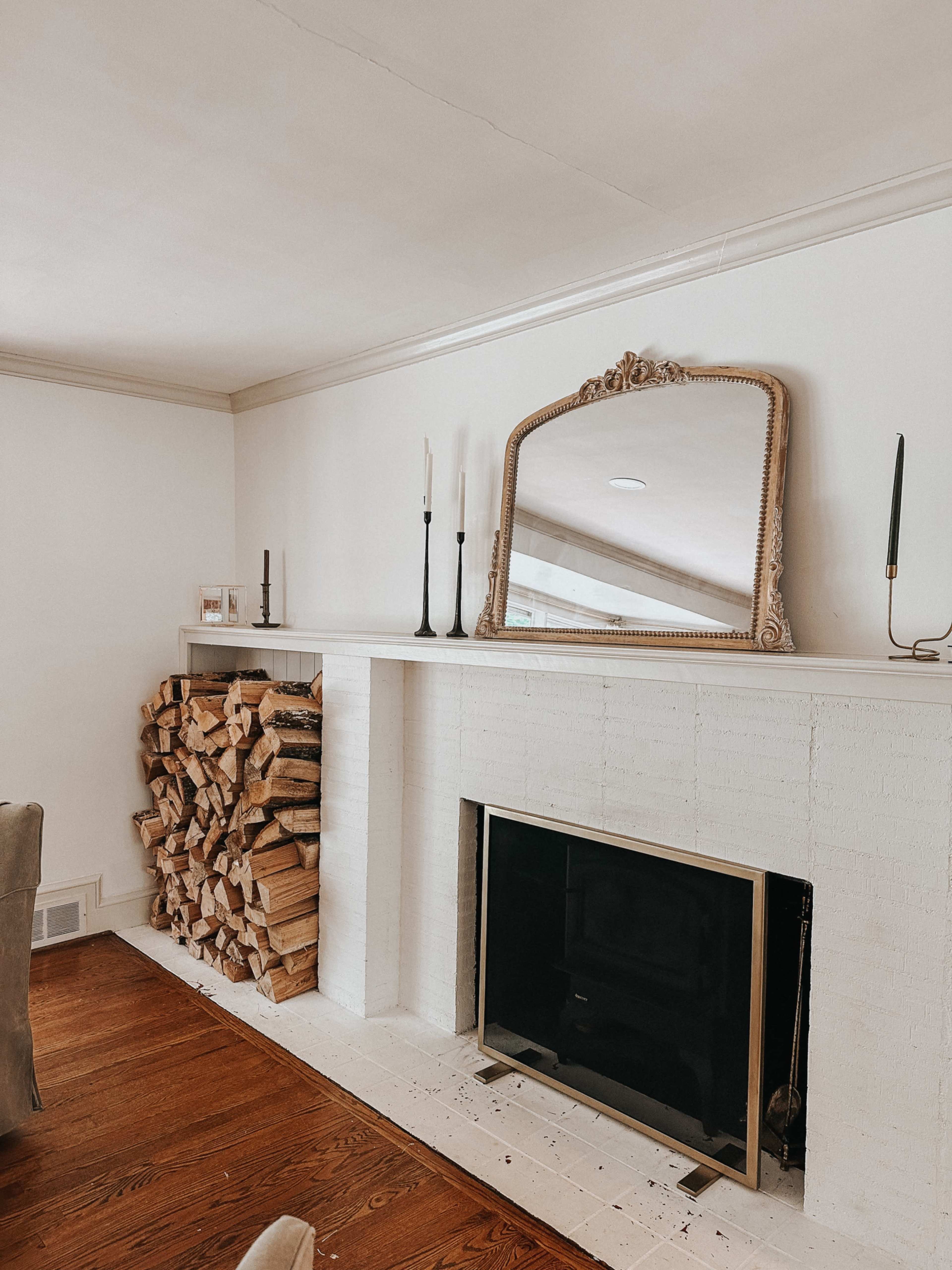 A living room with a stacked firewood display beside a white brick fireplace and a large mirror hanging above the mantle.