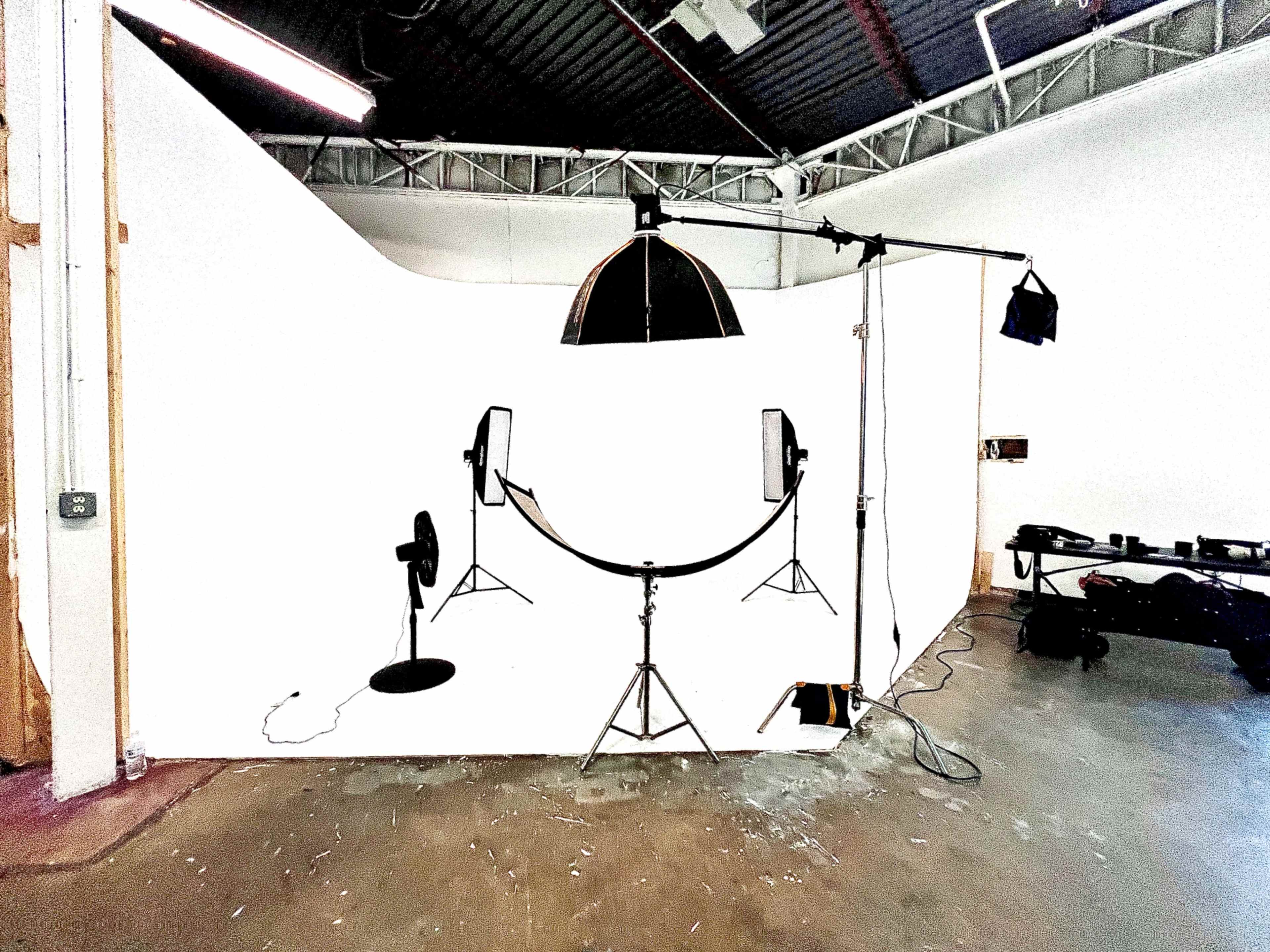 A photography studio setup with a curved white backdrop, two lighting stands, a fan, and various cables on a concrete floor.