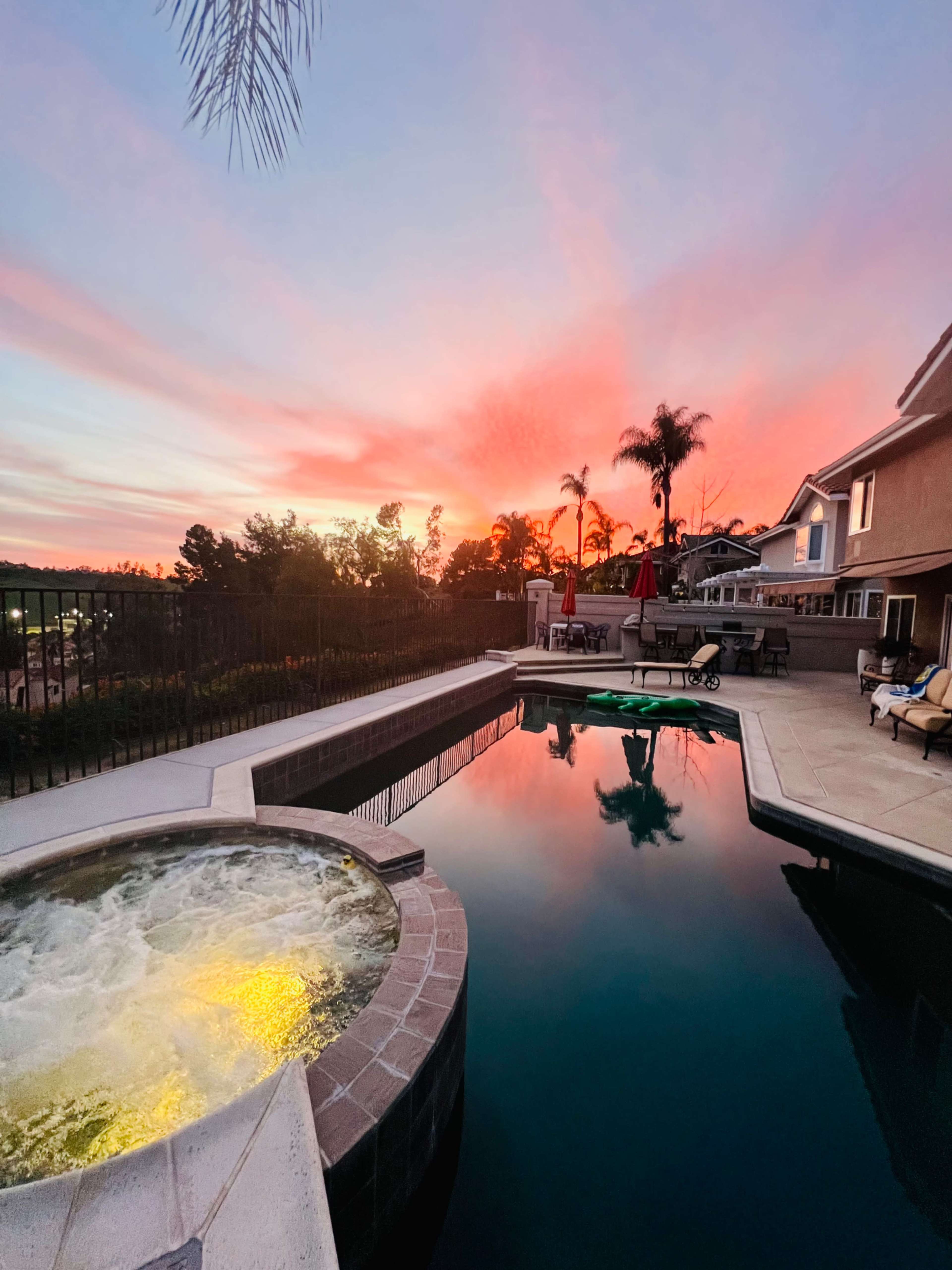 A swimming pool and hot tub are shown at sunset, with reflections of colorful clouds and palm trees in the water.