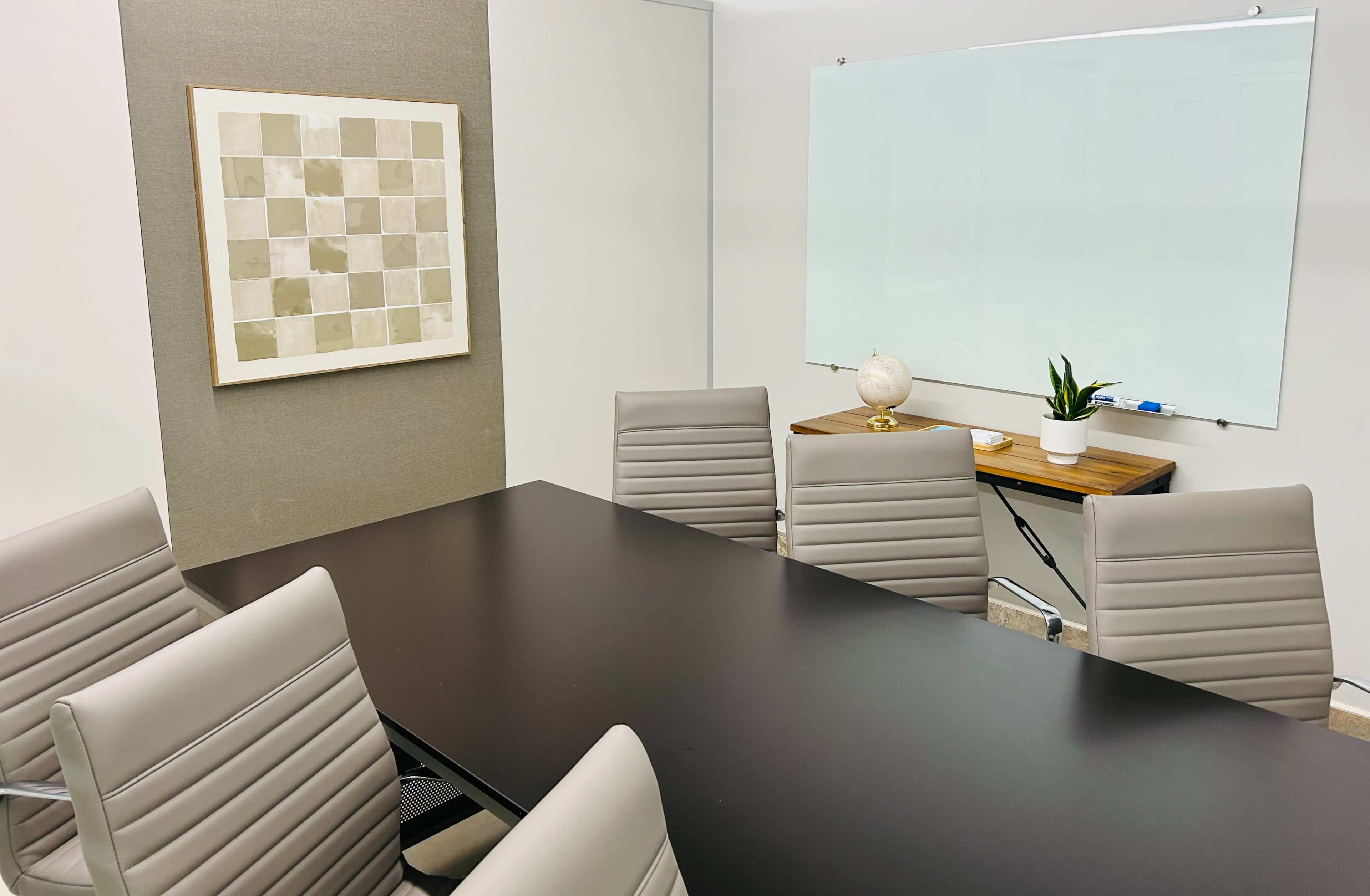 A modern conference room features a rectangular black table surrounded by light gray chairs, with a whiteboard and a decorative plant on a ledge.
