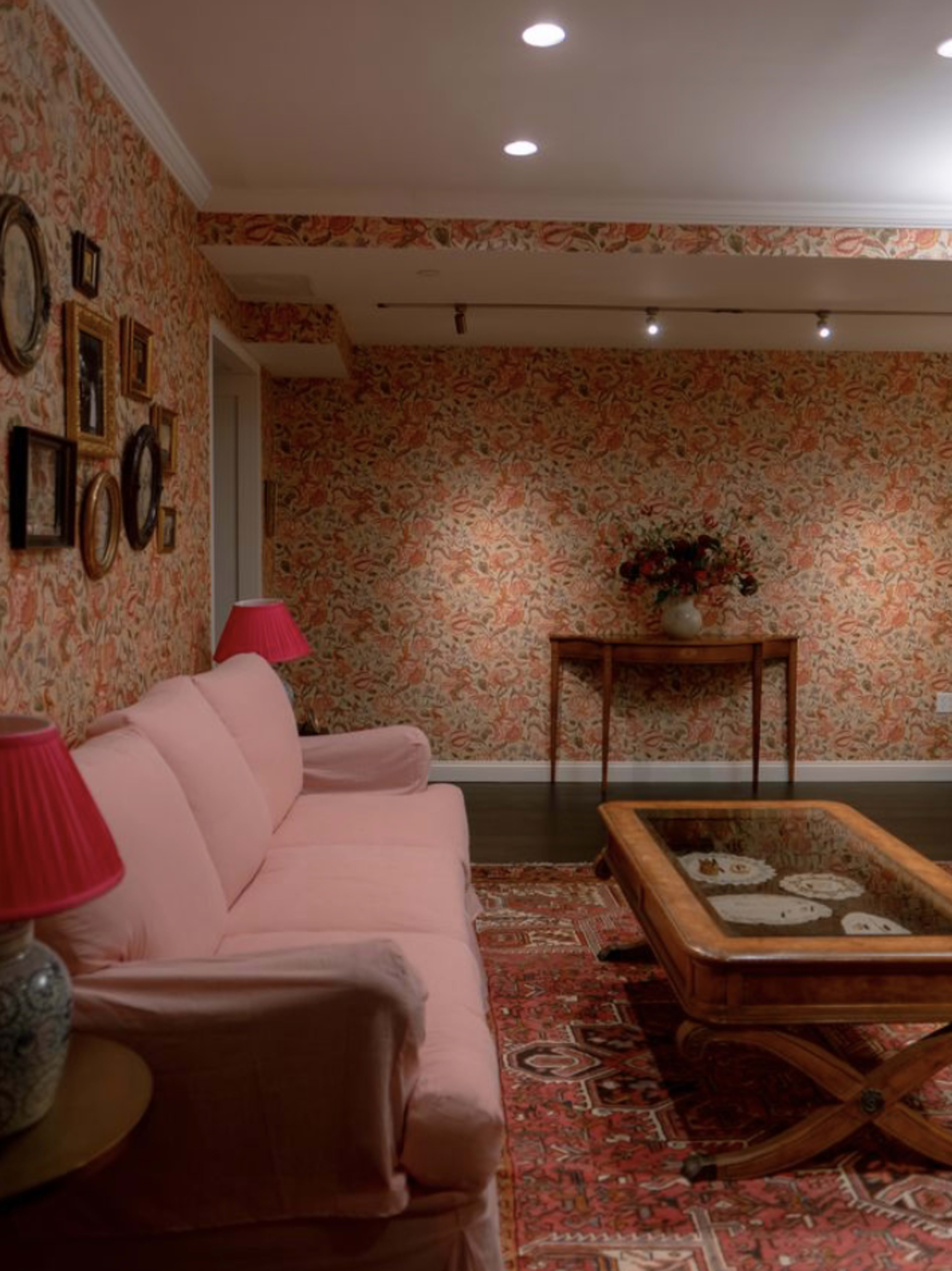 The image shows a cozy living room featuring a pink sofa, a wooden coffee table, and floral wallpaper with decorative frames on the walls.