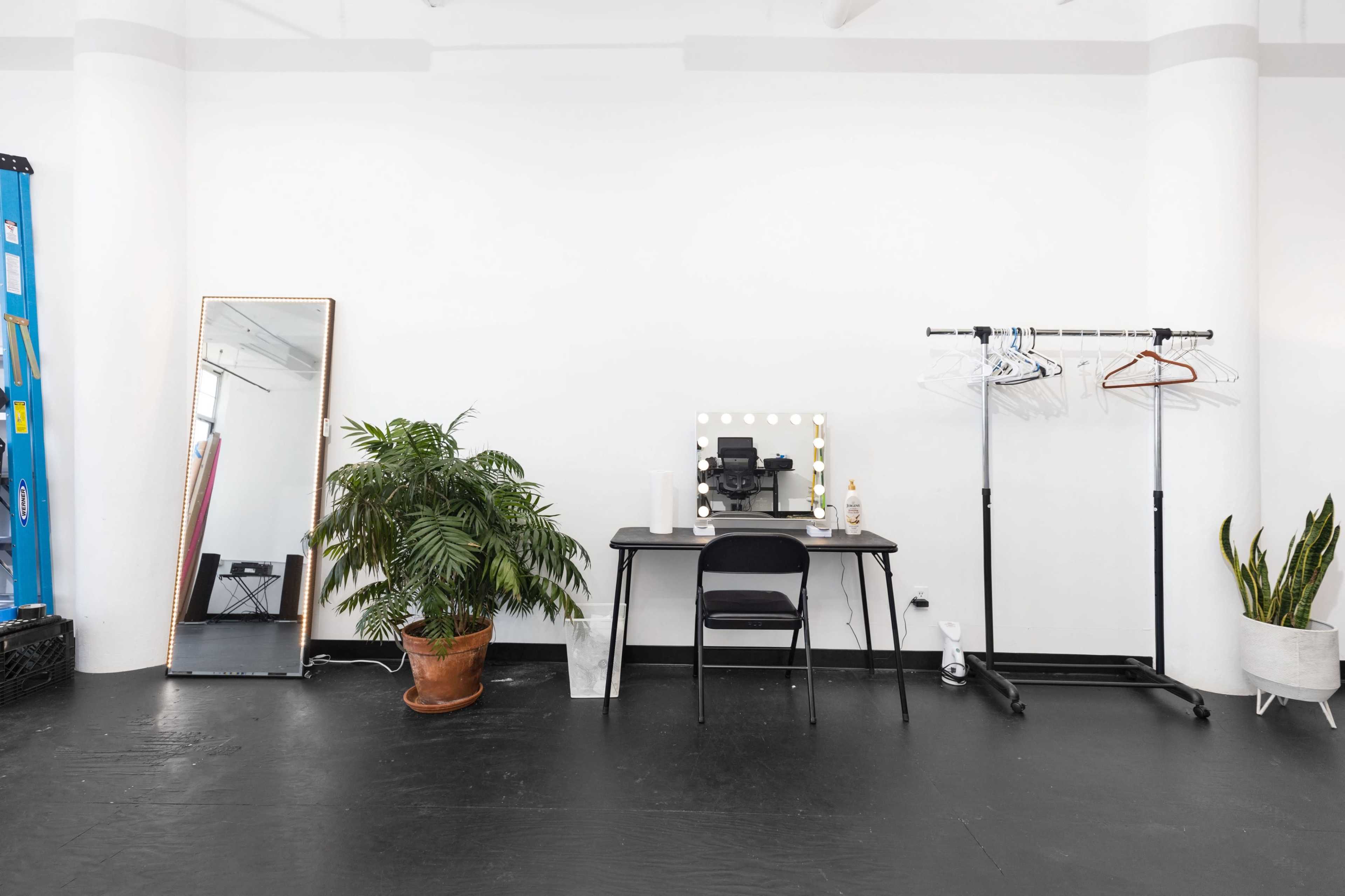 A minimalist room featuring a desk with a mirror and lights, a chair, a plant in a pot, a clothing rack, and a large mirror against a white wall.