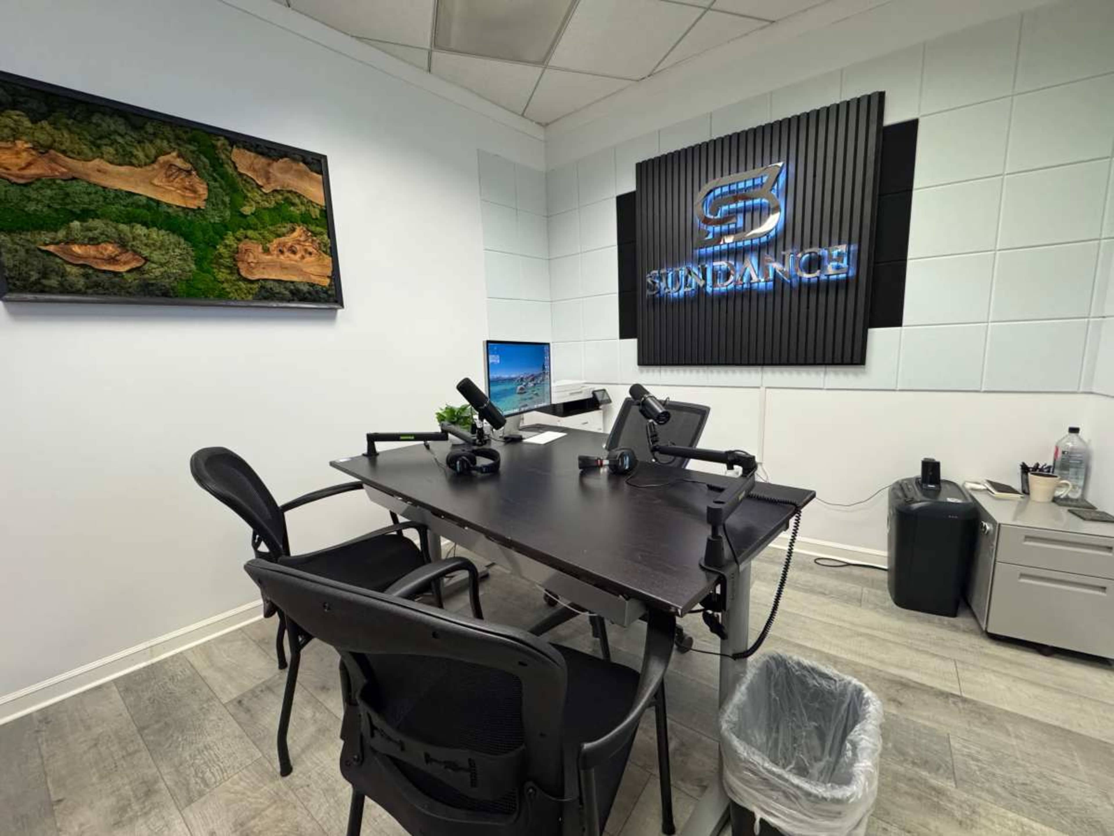 The image shows a modern office space with a black desk, two chairs, and equipment set up for video recording or streaming, including microphones and computers, against a backdrop featuring a company logo.