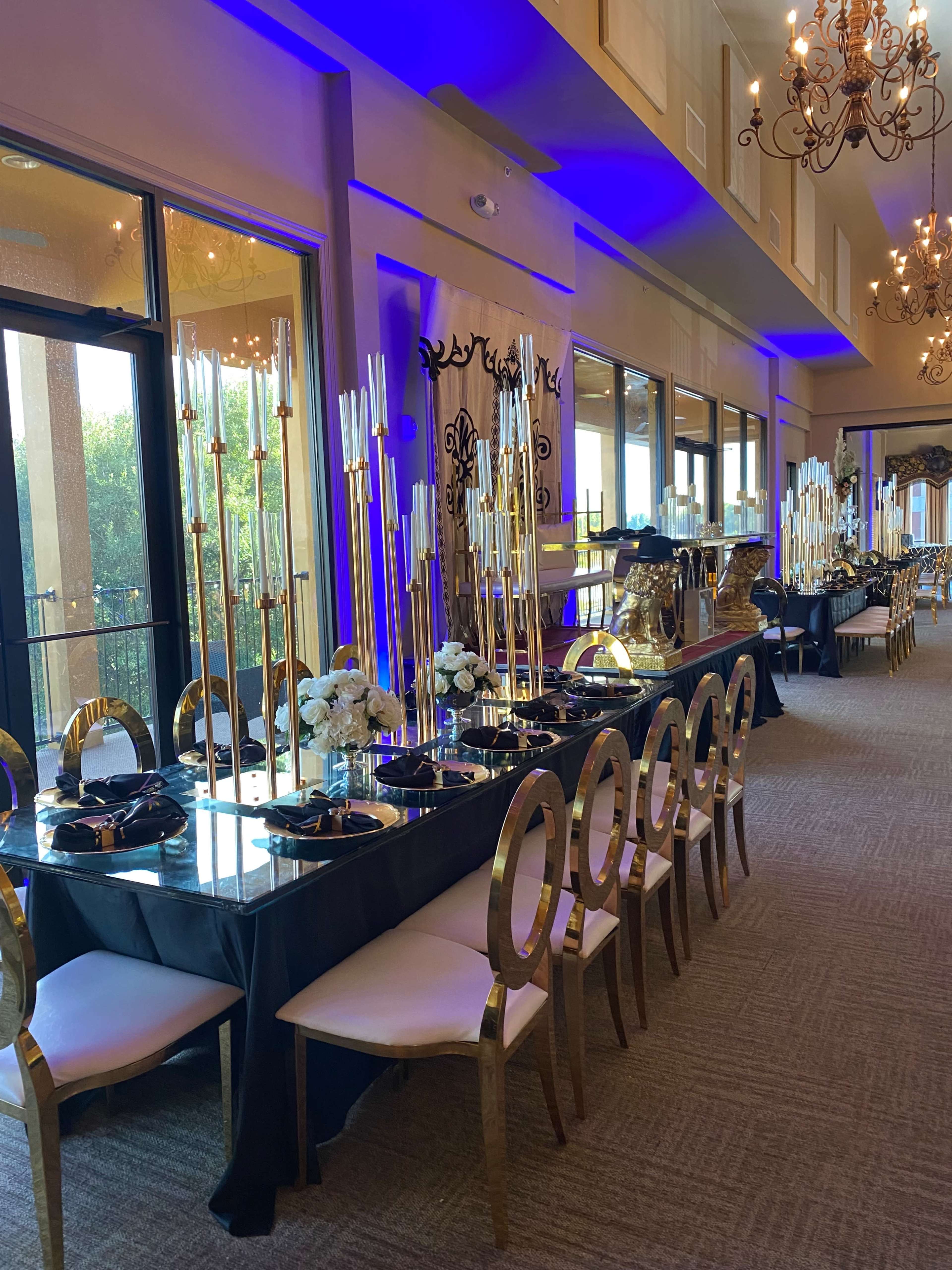 A dining area is set with elegant tables featuring tall candlesticks and floral arrangements, illuminated by soft blue lighting.