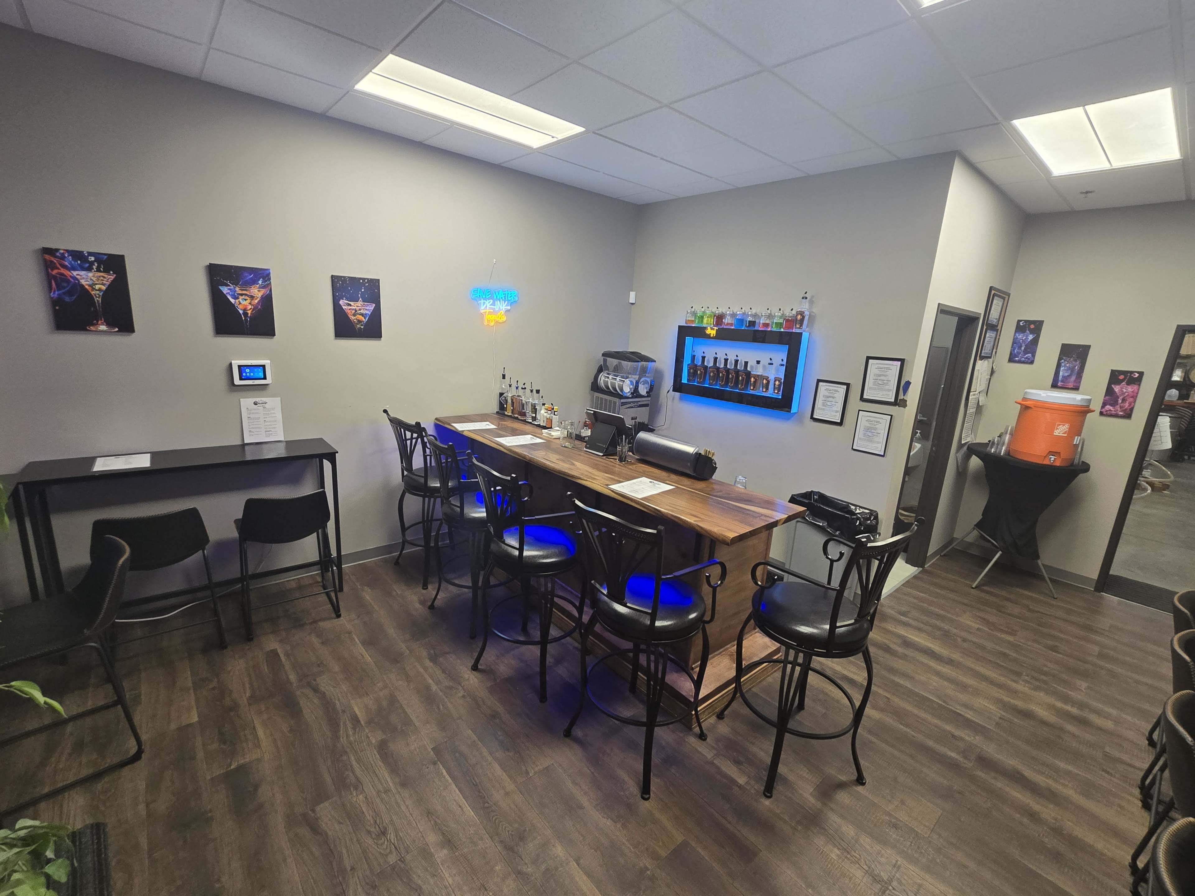 The image shows a small bar area with a counter, several chairs, and a wall displaying colorful artwork and a neon sign.