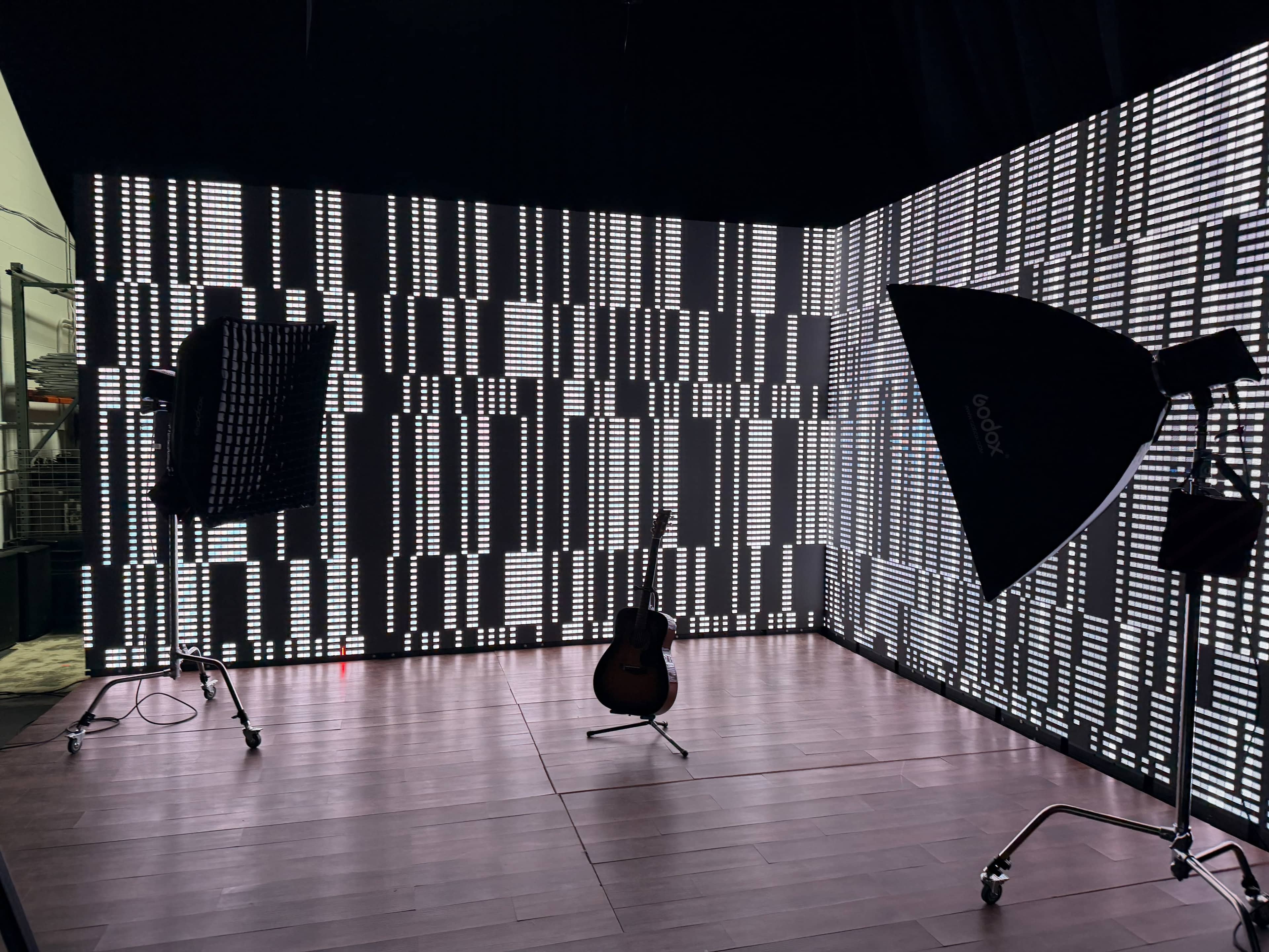 The image shows a dimly lit studio space with a wooden floor and walls covered in light strips, featuring a guitar placed in the center and professional lighting equipment set up nearby.