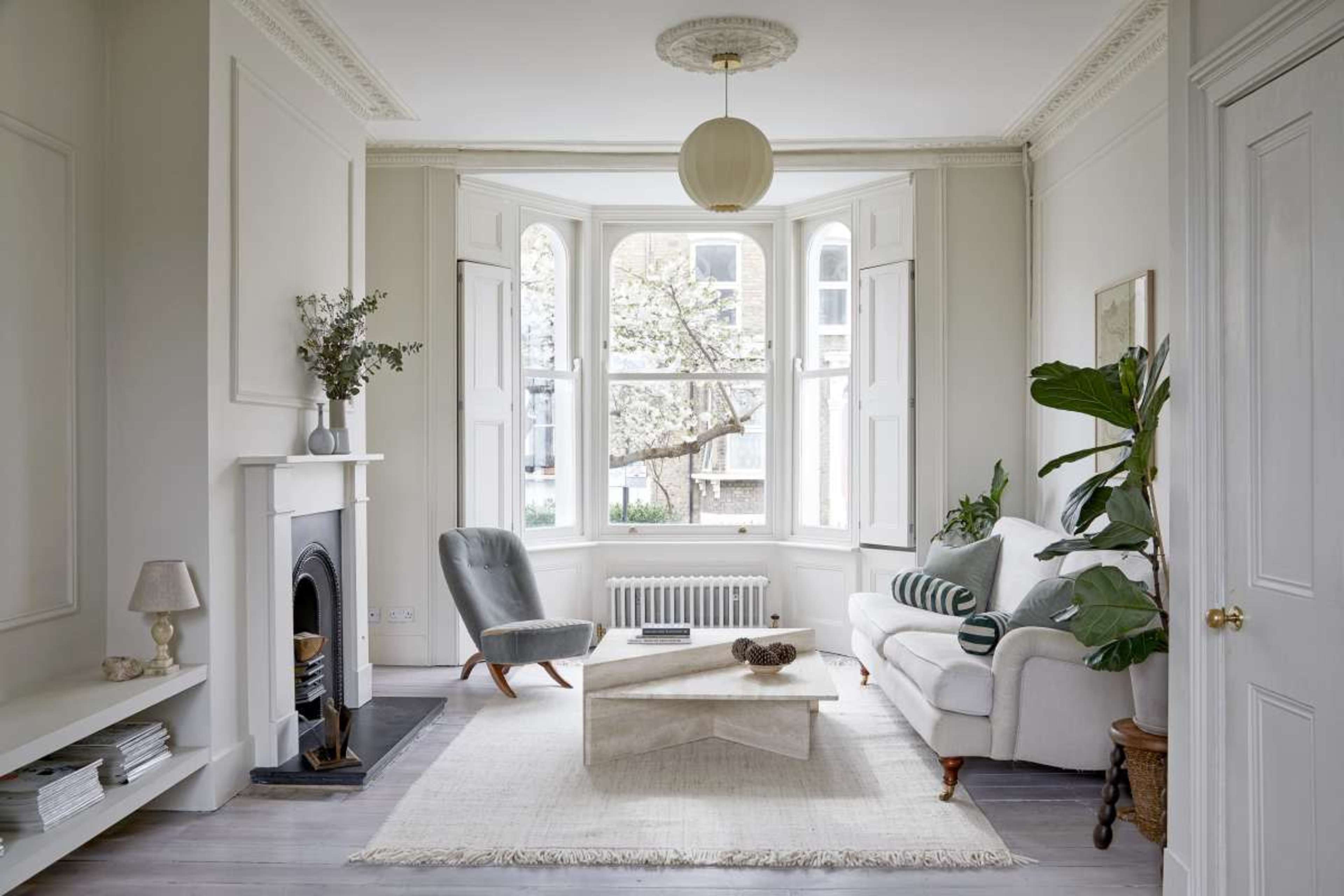 The image shows a bright, minimalist living room featuring a white sofa, a coffee table, and large windows with a view of trees outside.