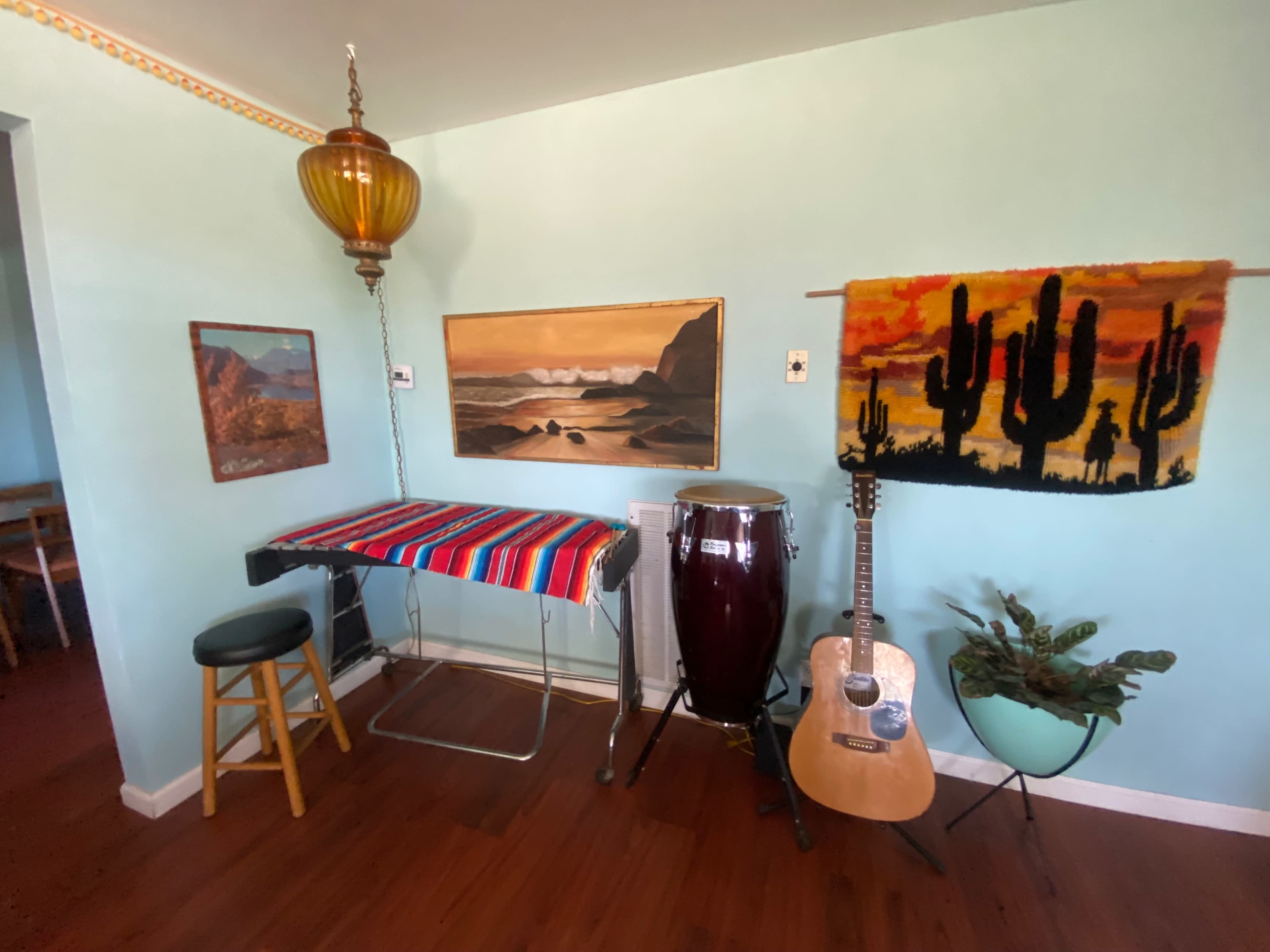 The image shows a corner of a room with a colorful striped table cover, a wooden stool, a conga drum, an acoustic guitar, a cactus-themed wall hanging, and framed artwork on the walls.