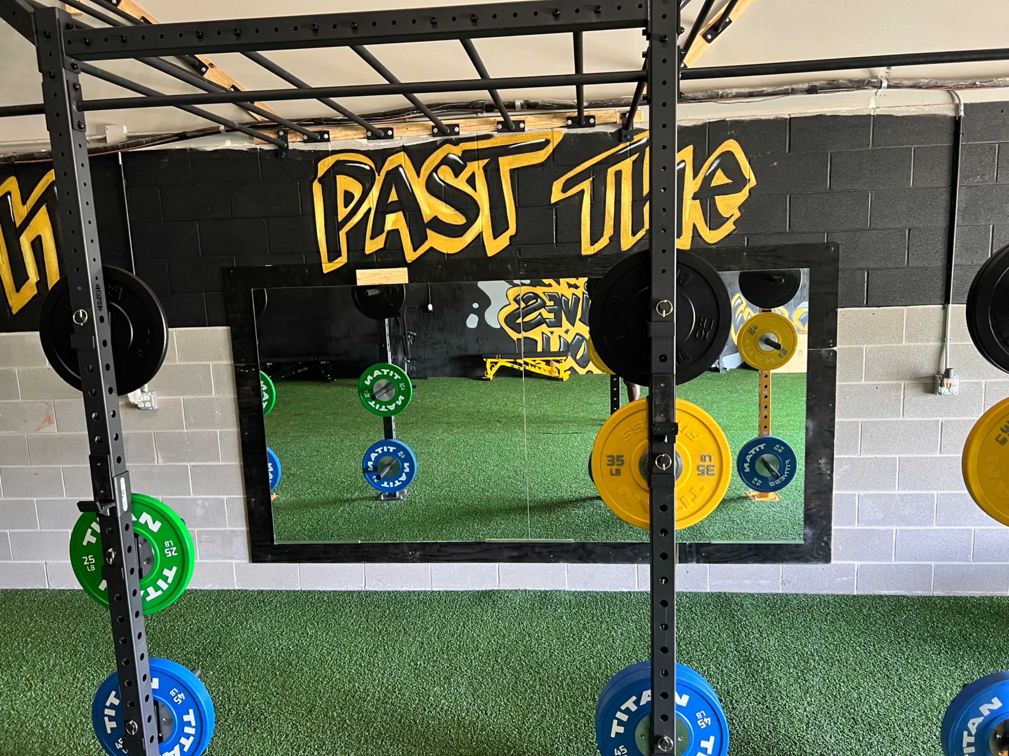 A weightlifting area with a mirror mounted on a wall, surrounded by colored weight plates on a rack and artificial turf flooring.