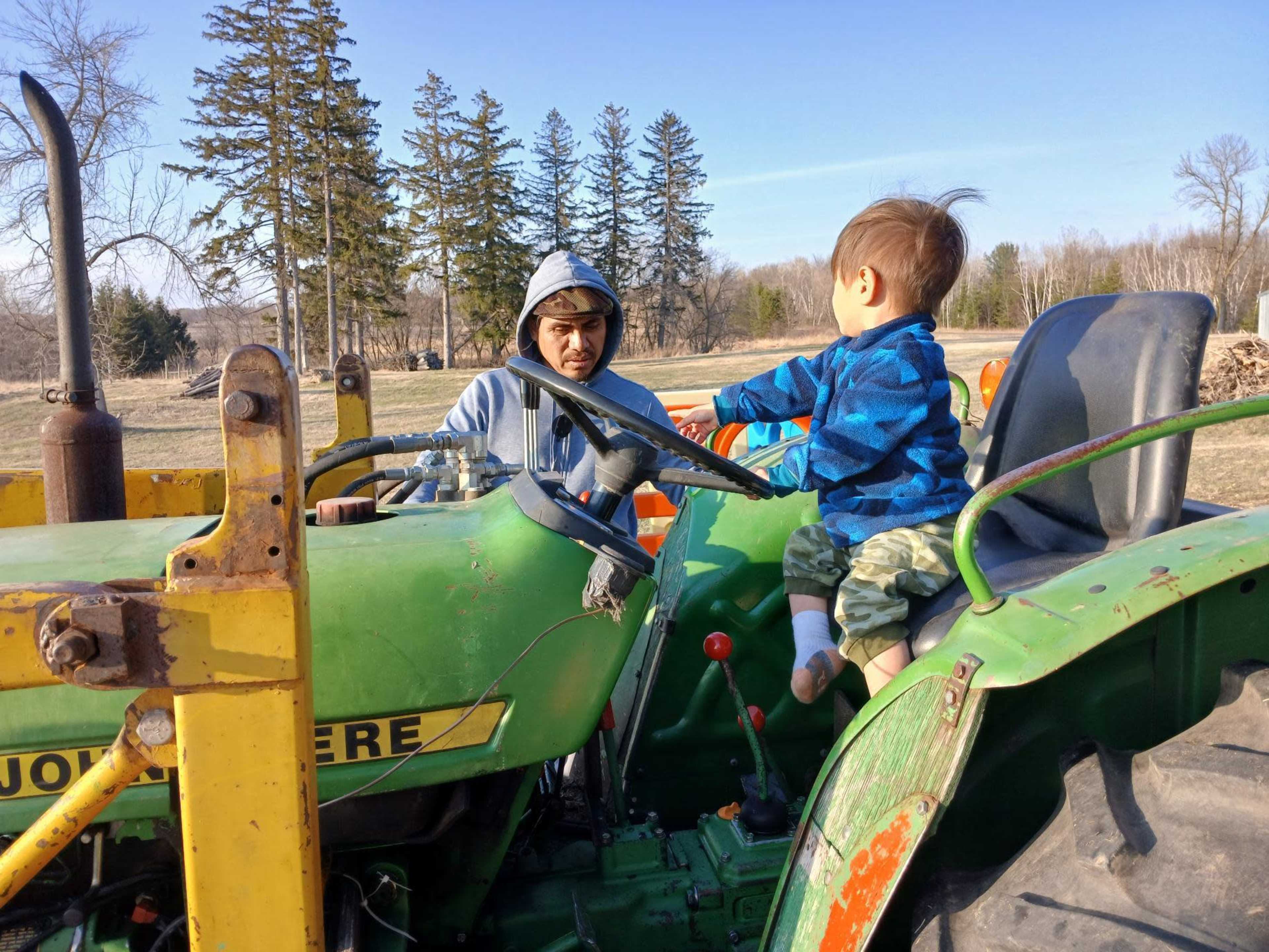 A young child sits on a tractor's seat while an older boy looks on in a rural setting.