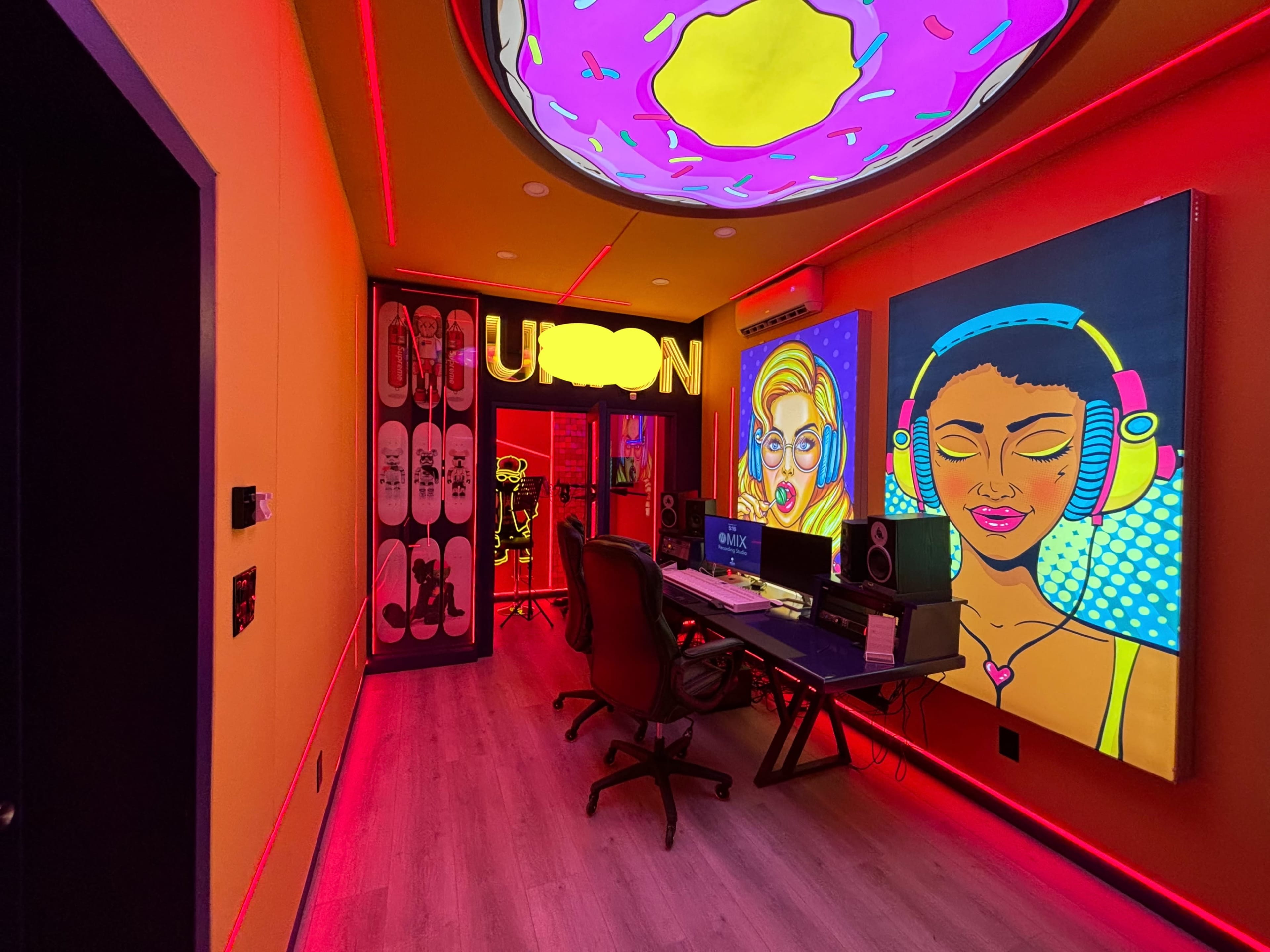 A brightly colored music studio featuring neon artwork, a desk with speakers, and skateboards mounted on the wall.