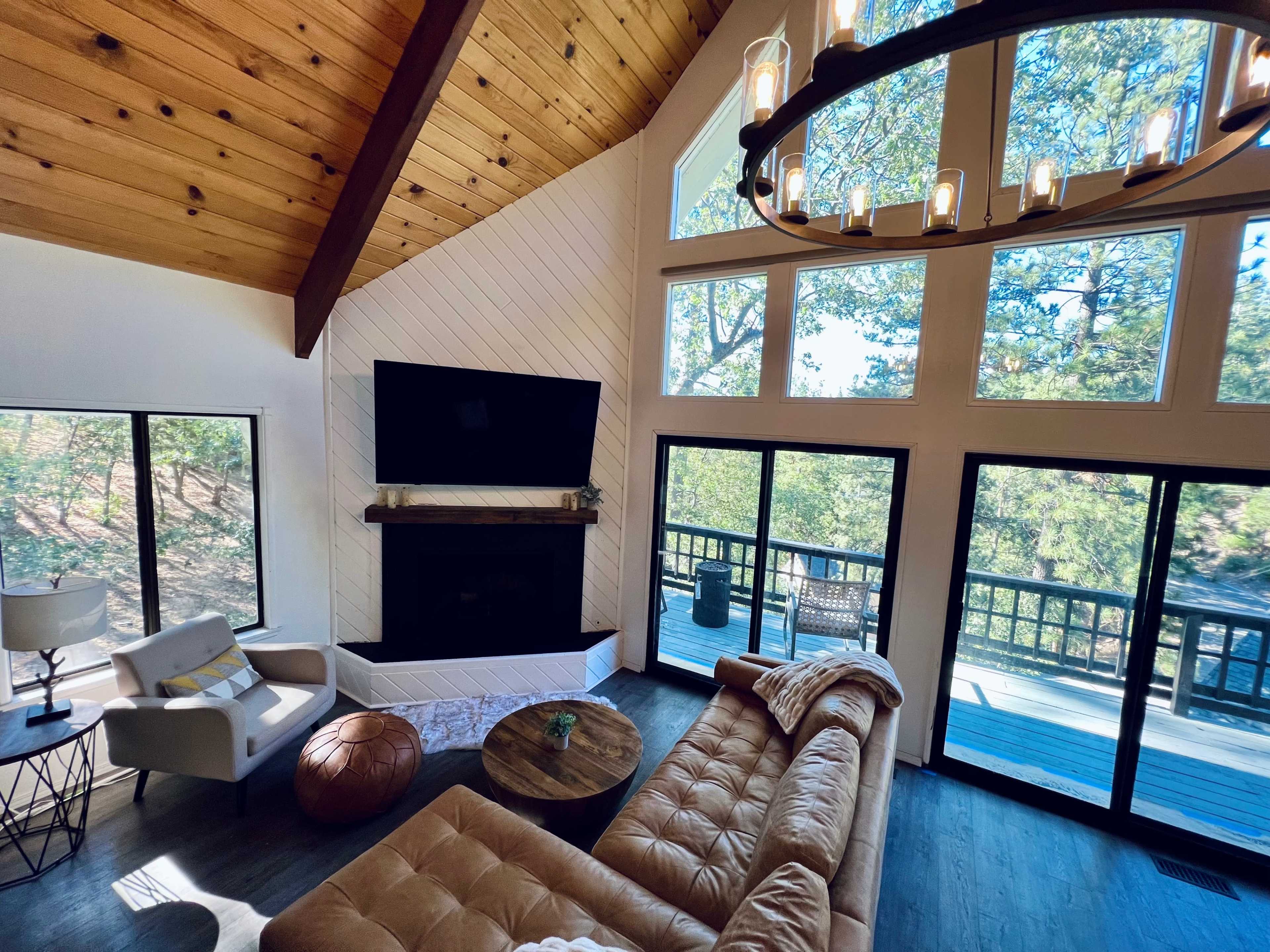The image shows a modern living room with a large sectional sofa, a fireplace, and large windows that overlook a wooded area.