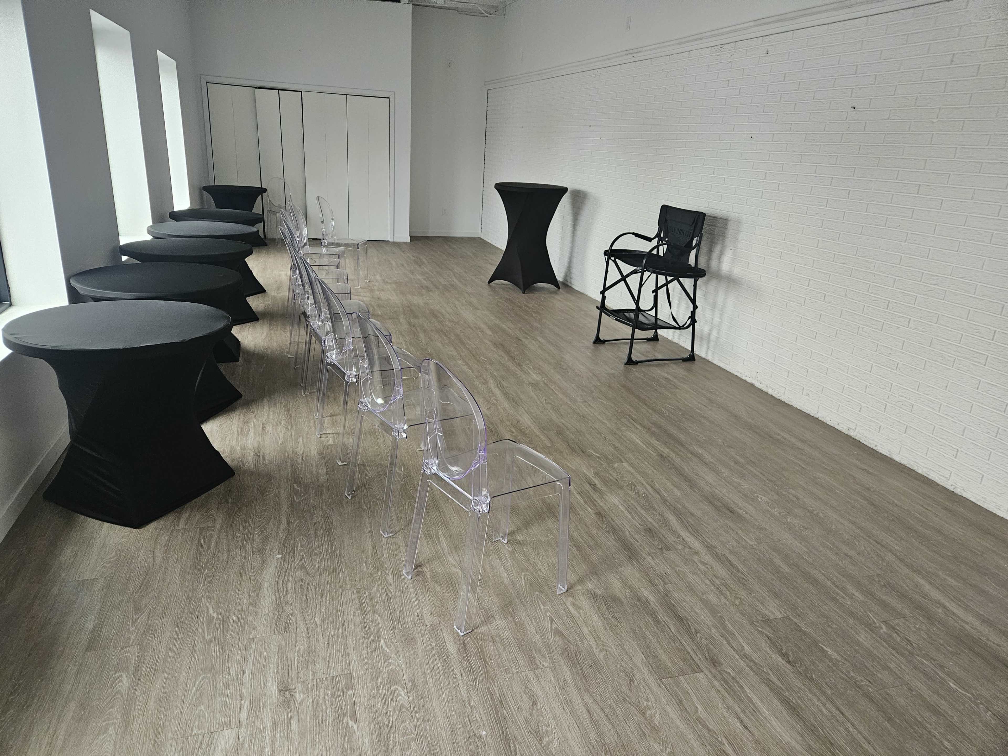 The image shows a spacious, empty room with a row of transparent chairs and several cocktail tables, alongside a director's chair and a small table set against a white brick wall.