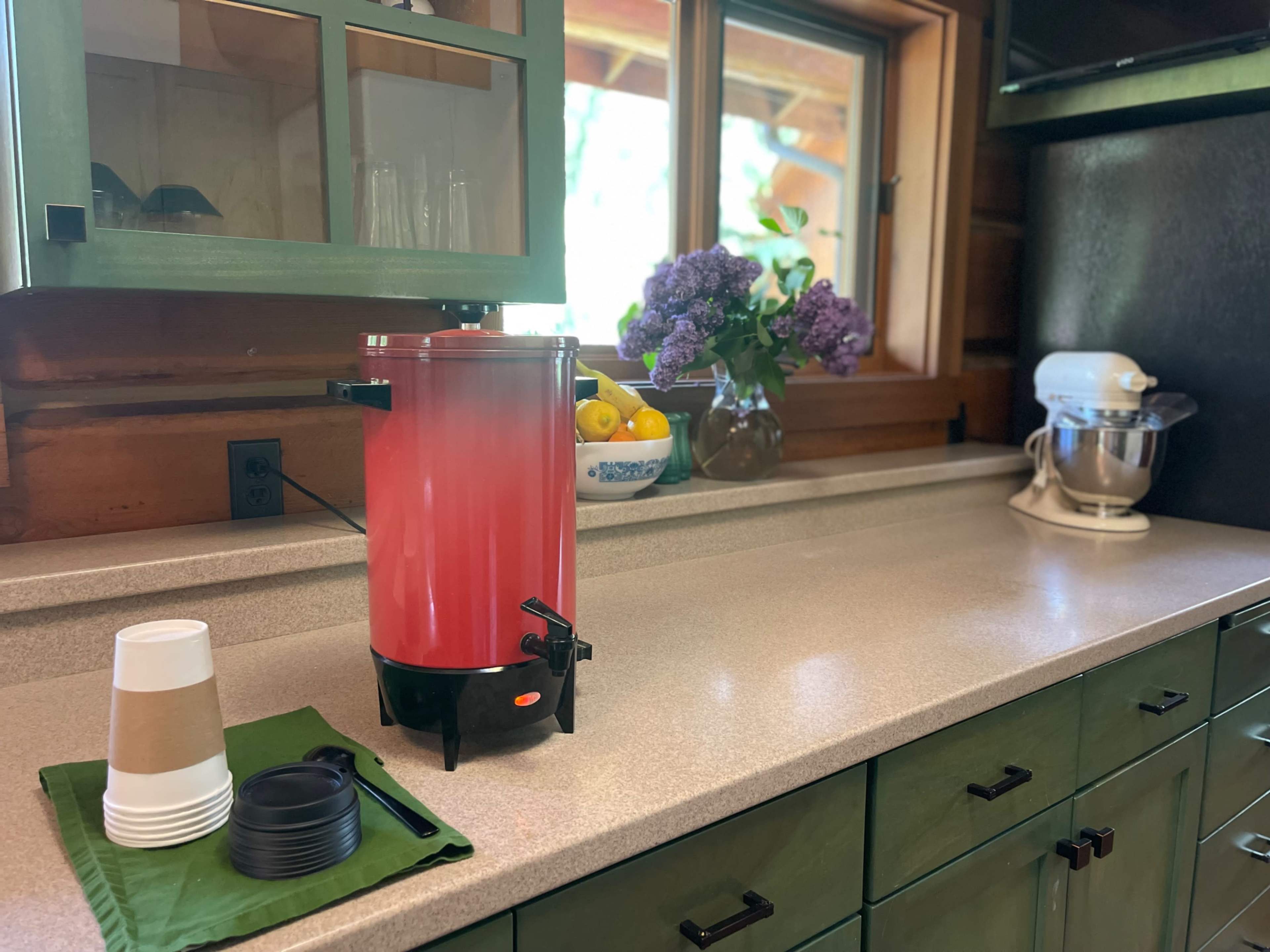 A red beverage dispenser sits on a countertop next to a bowl of fruit and a bouquet of flowers in a kitchen with wooden cabinetry.