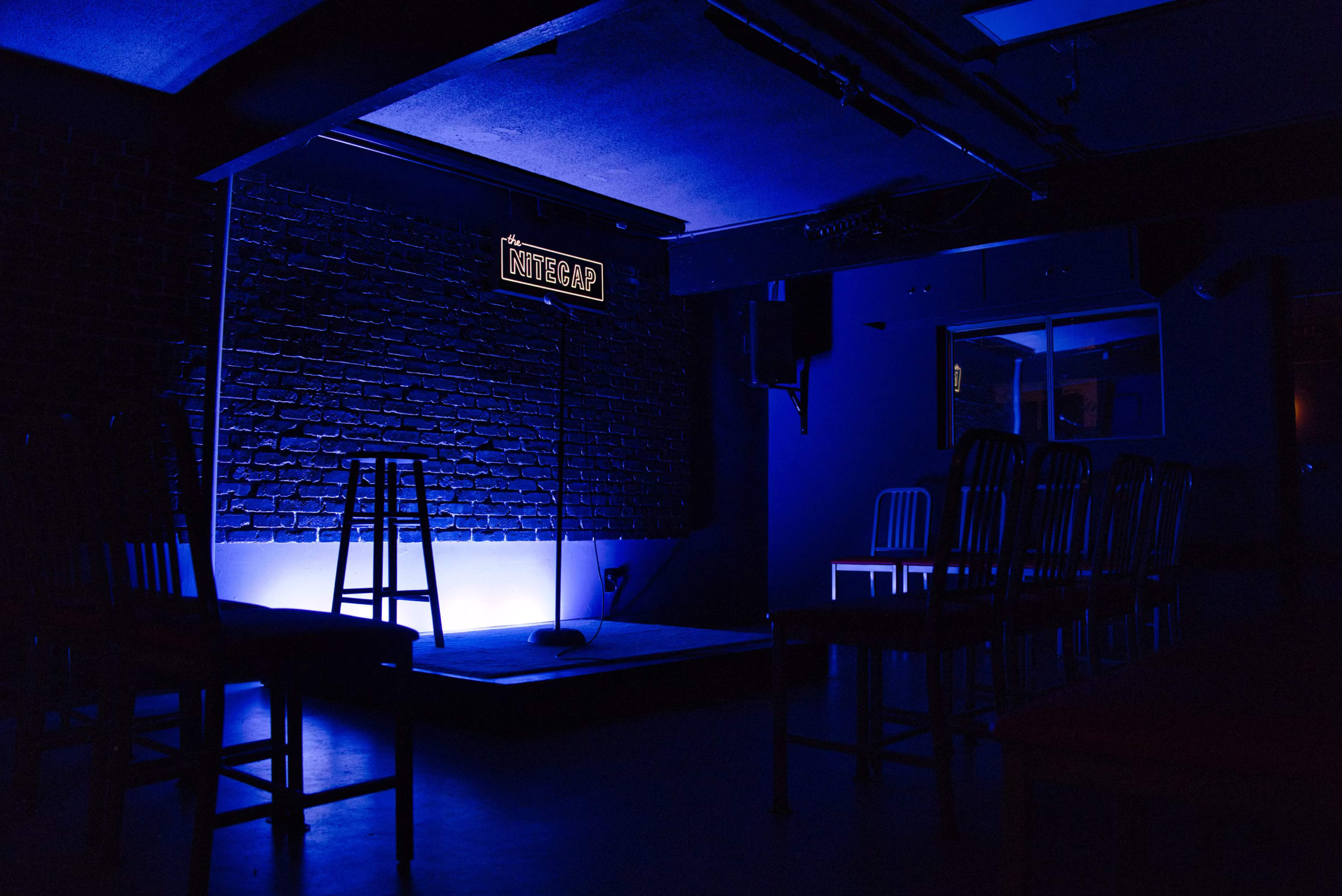 A dimly lit comedy club stage features a microphone stand and a stool, with blue lighting illuminating the area.