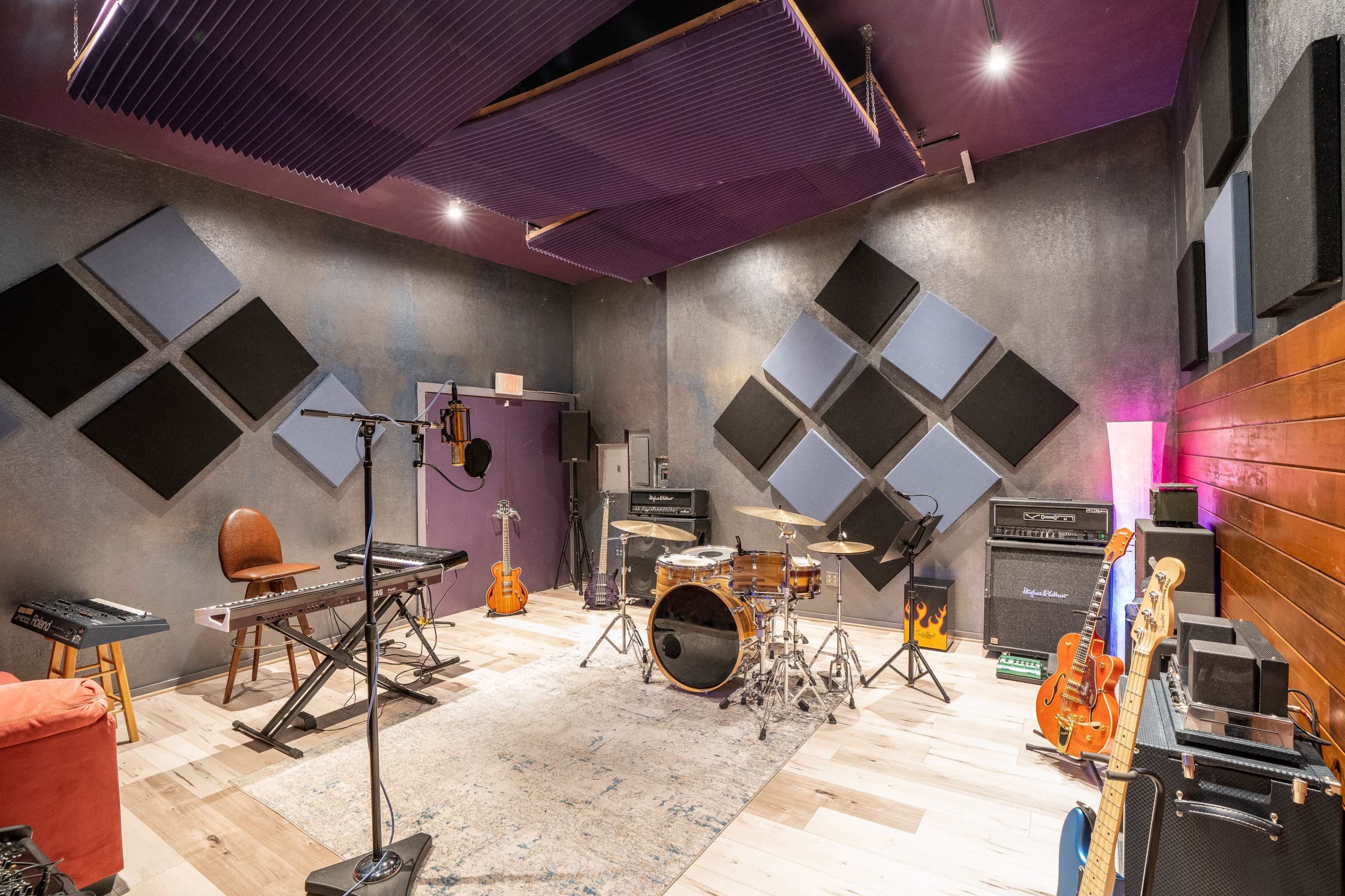 A music recording studio featuring a drum set, keyboard, guitar amplifiers, and various instruments arranged around a rug on a wooden floor.