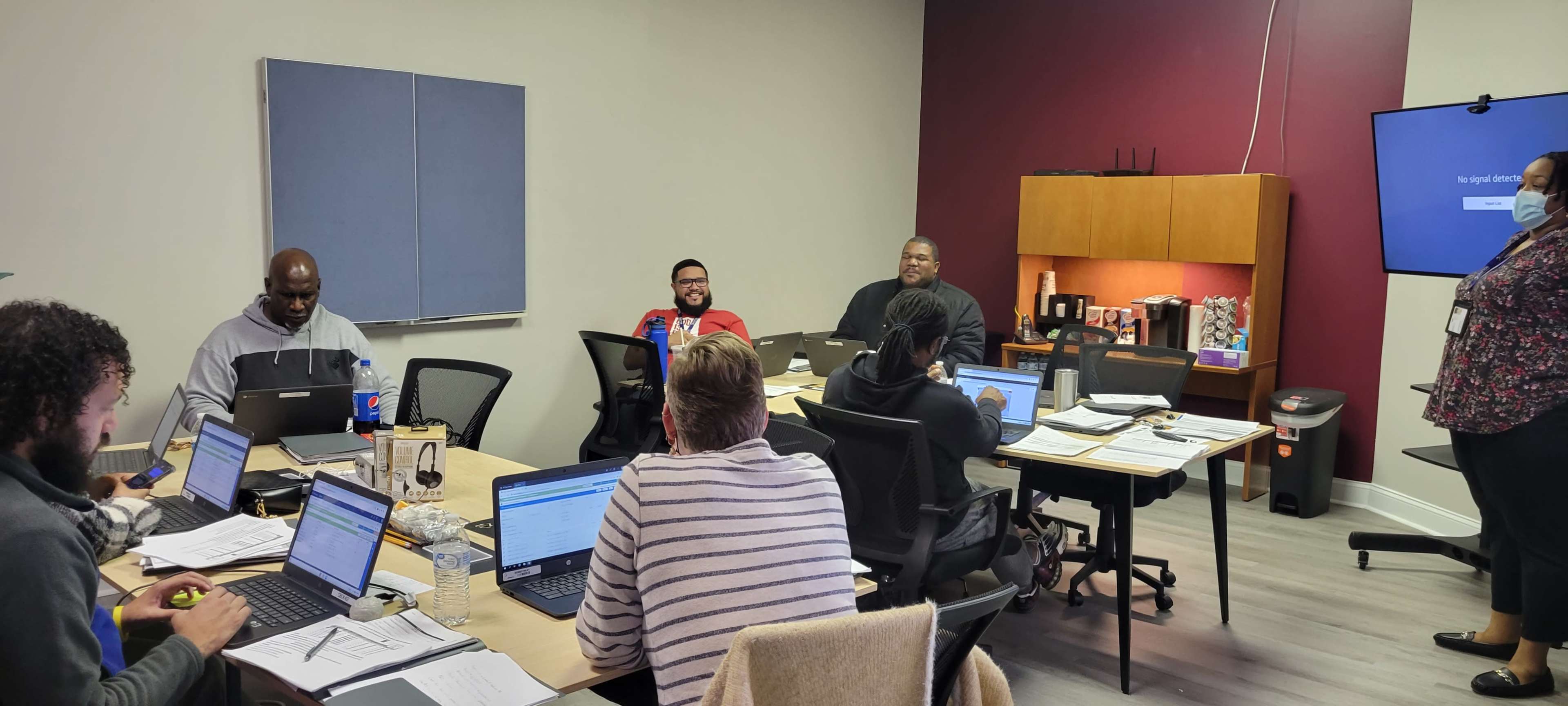 A group of eight people are seated around a conference table in an office, working on laptops while engaging in a discussion.