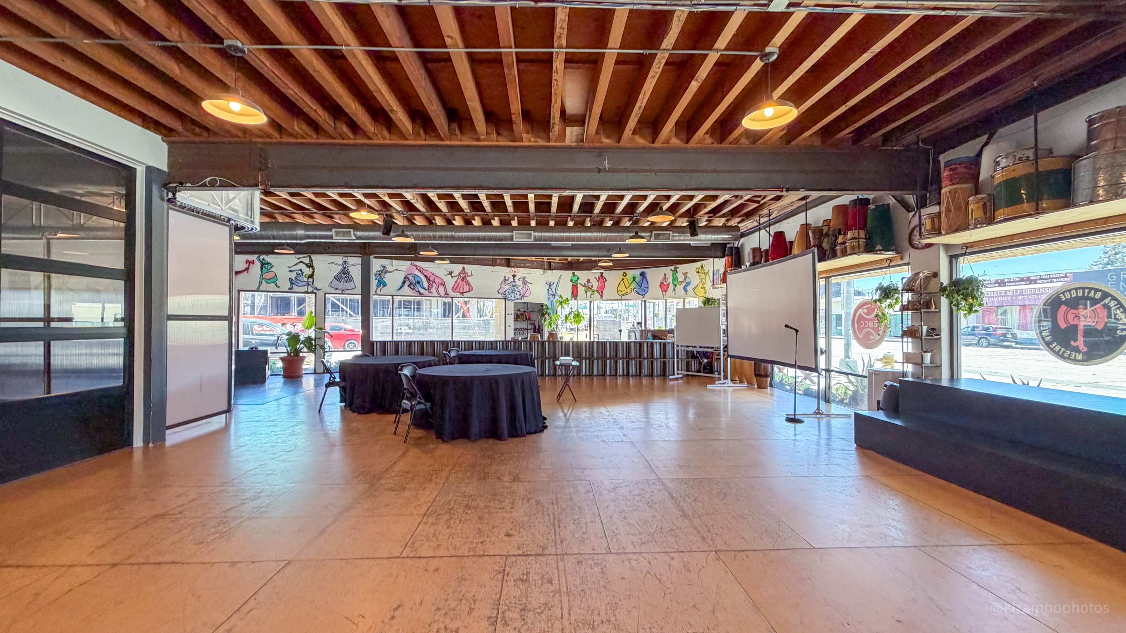 The image shows a spacious event venue with wooden ceilings, empty tables, and colorful wall murals.