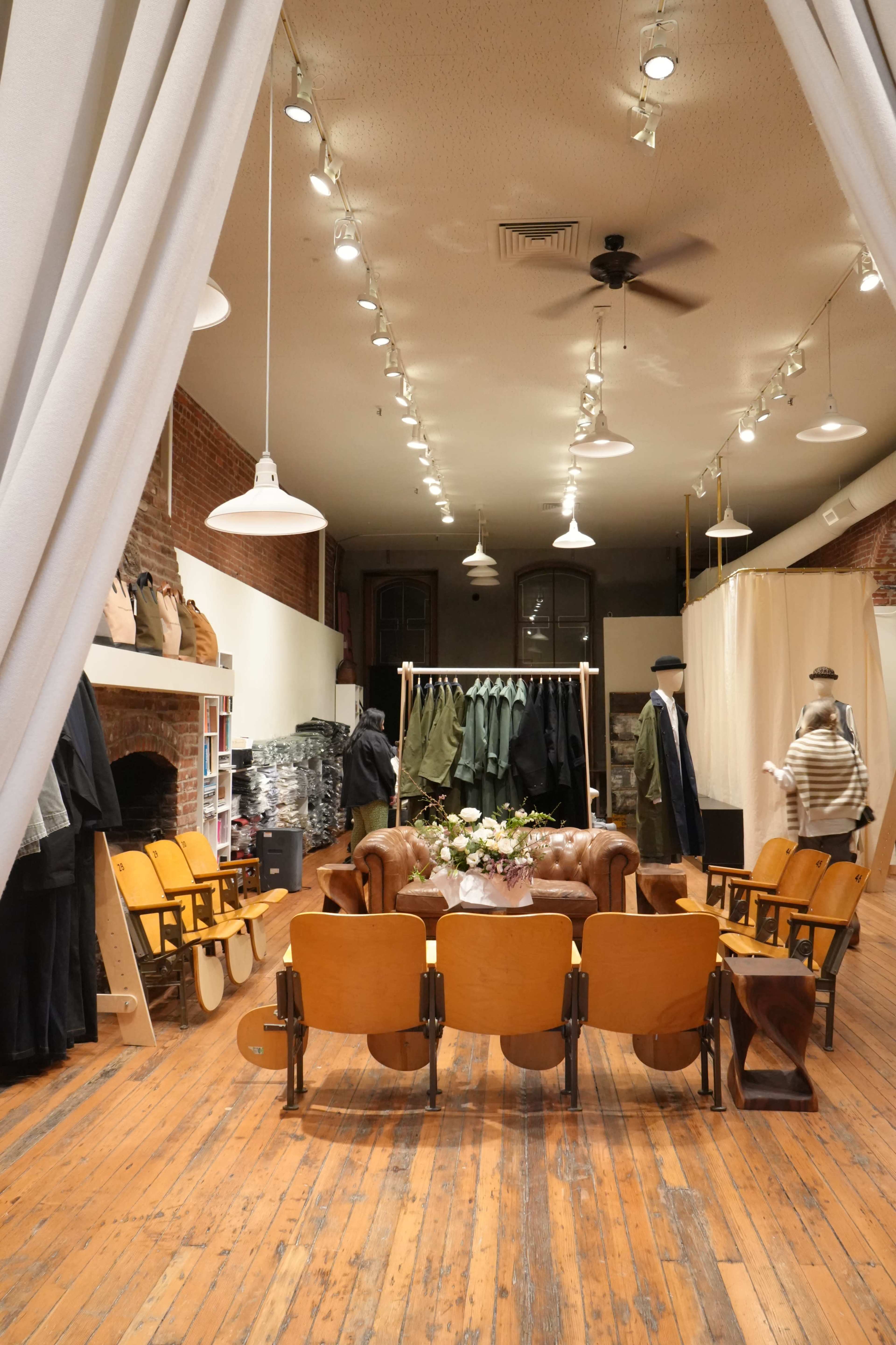 The image shows a retail space with a rustic wooden floor, featuring a circular arrangement of chairs, a table with flowers, and racks of clothing displayed along the walls.