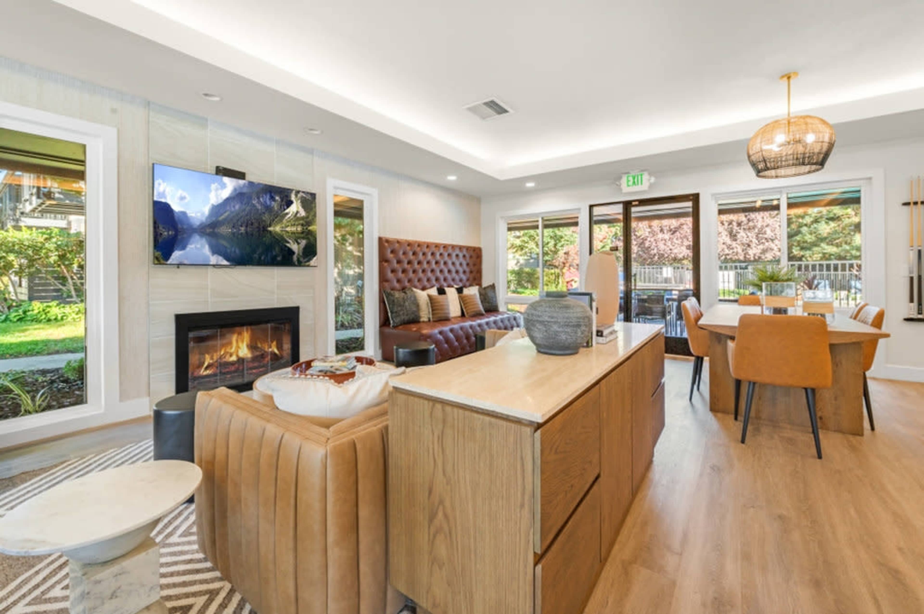The image shows a modern living room with a fireplace, a TV mounted on the wall, a dining area, and large windows letting in natural light.