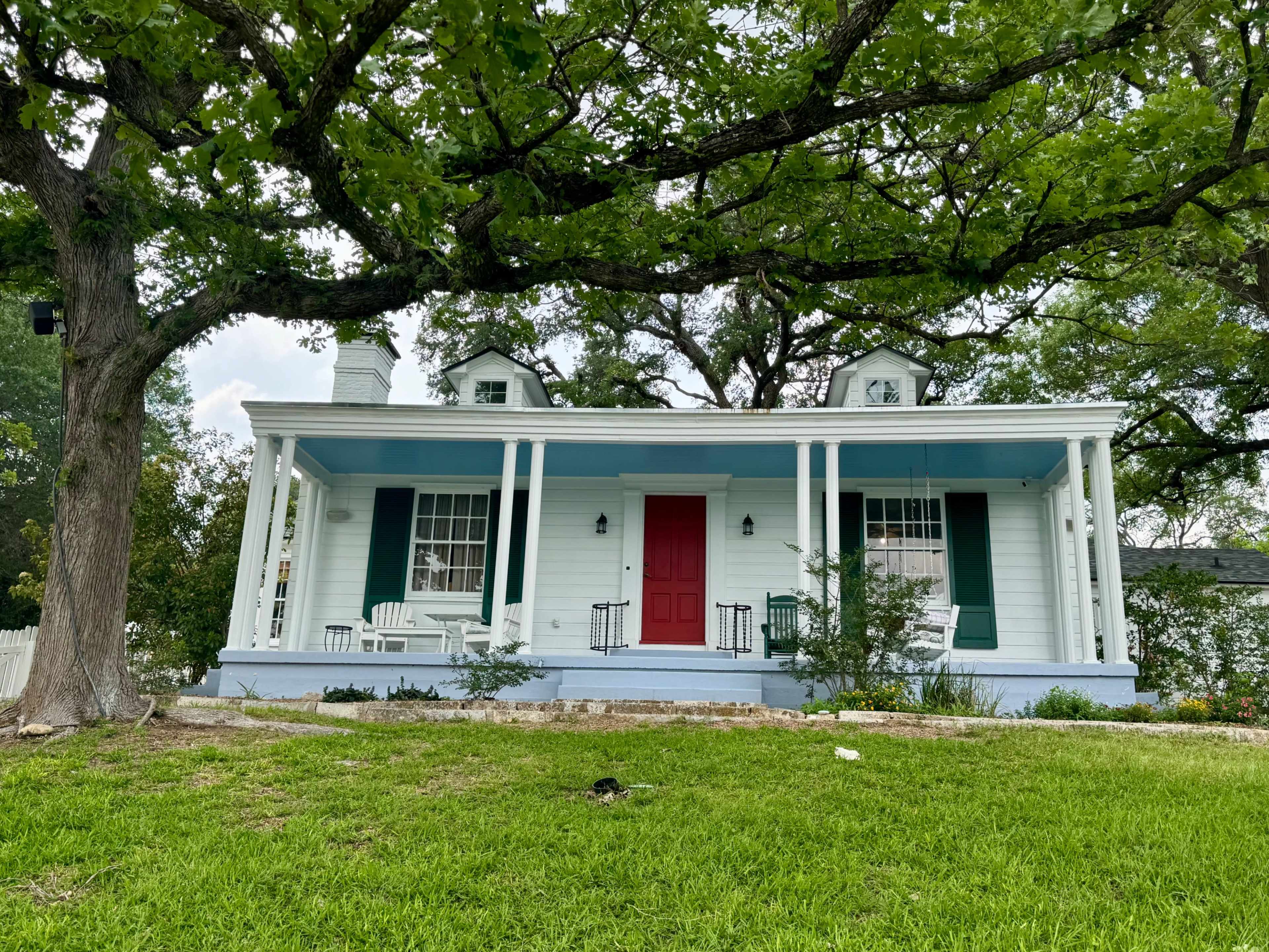 The image shows a light blue and white house with a red front door, flanked by two small gabled windows and surrounded by a grassy yard and large trees.