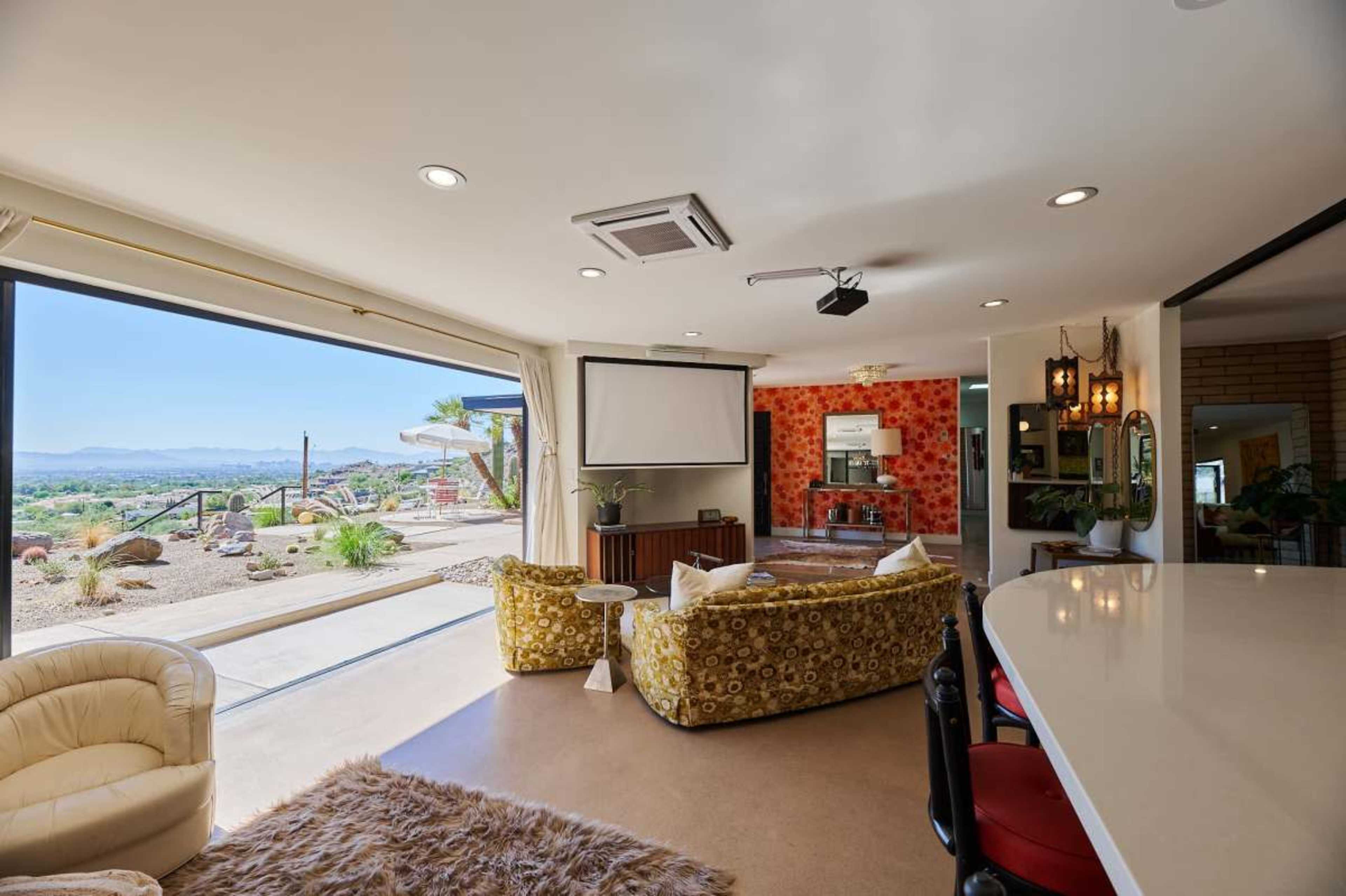 The spacious living area features large glass doors opening to a mountain view, with modern furniture and a projector screen mounted on the ceiling.
