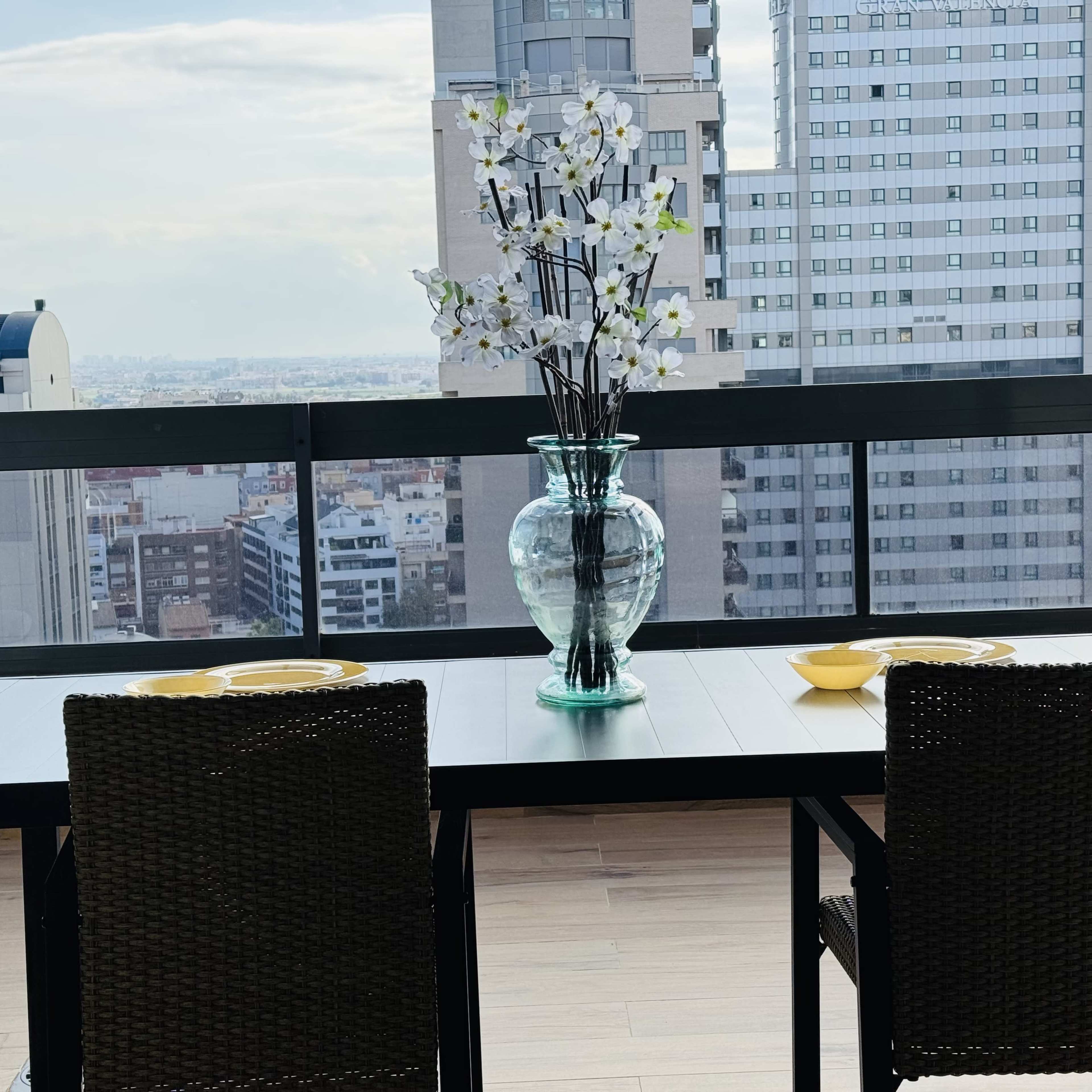 A dining table with two chairs and a large vase filled with flowers sits on a balcony overlooking a city skyline.