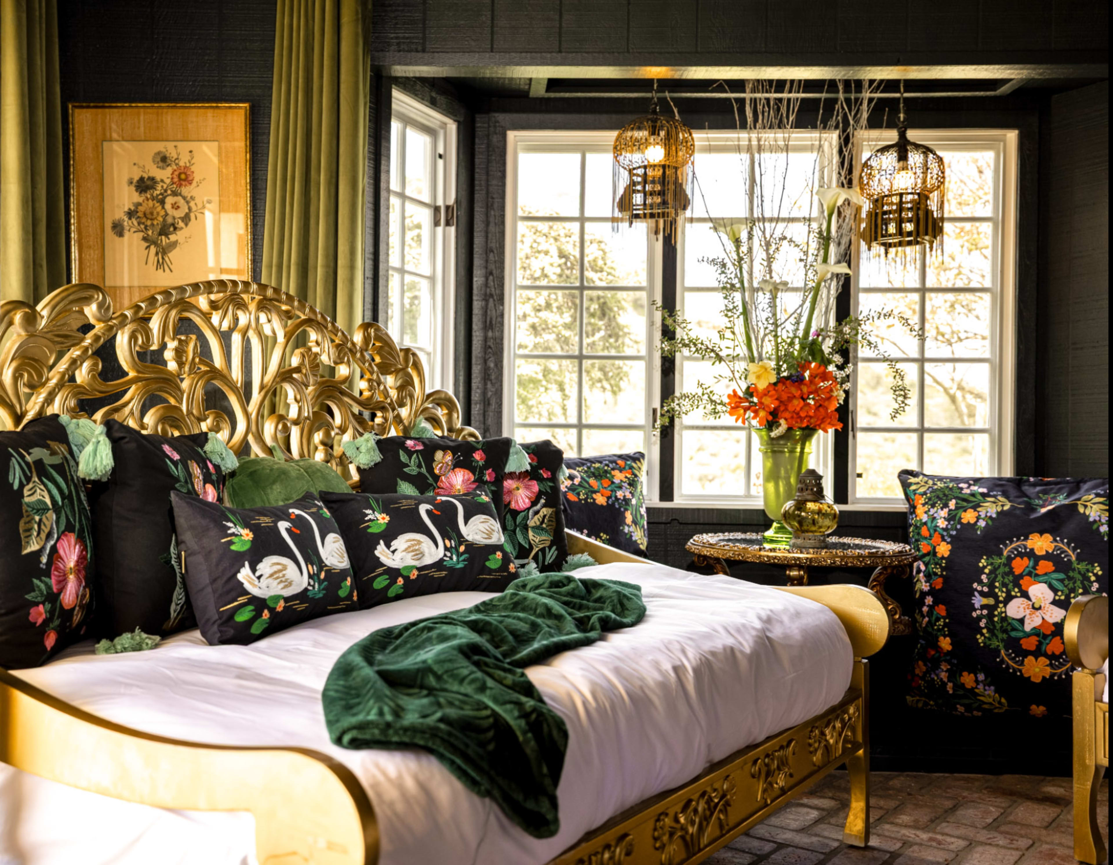The room features a gold ornate bed with dark floral decorative pillows, a vase of flowers on a side table, and large windows allowing natural light to enter.