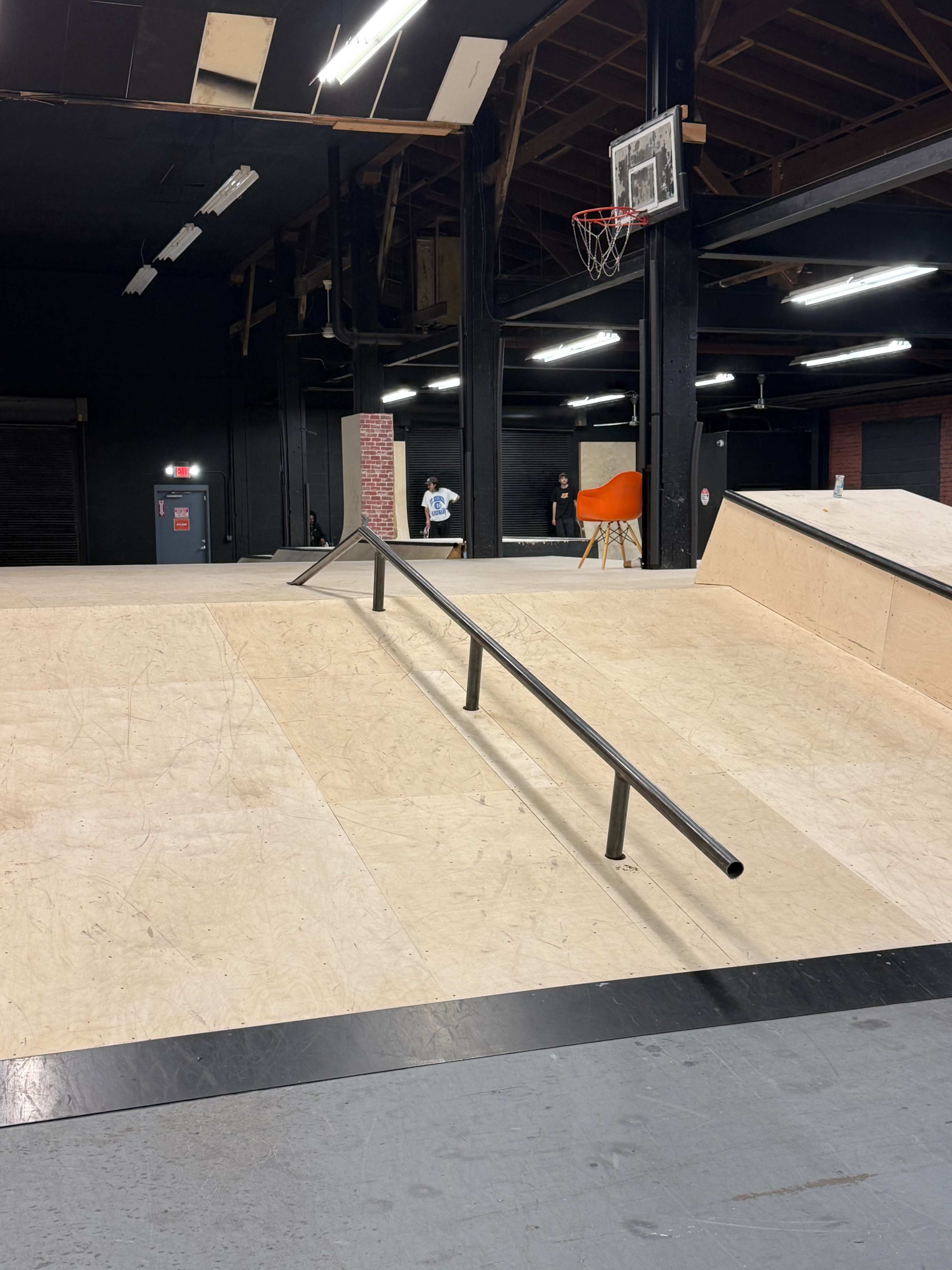 The image shows a skate park interior with a wooden ramp and a metal rail, along with a basketball hoop in the background.