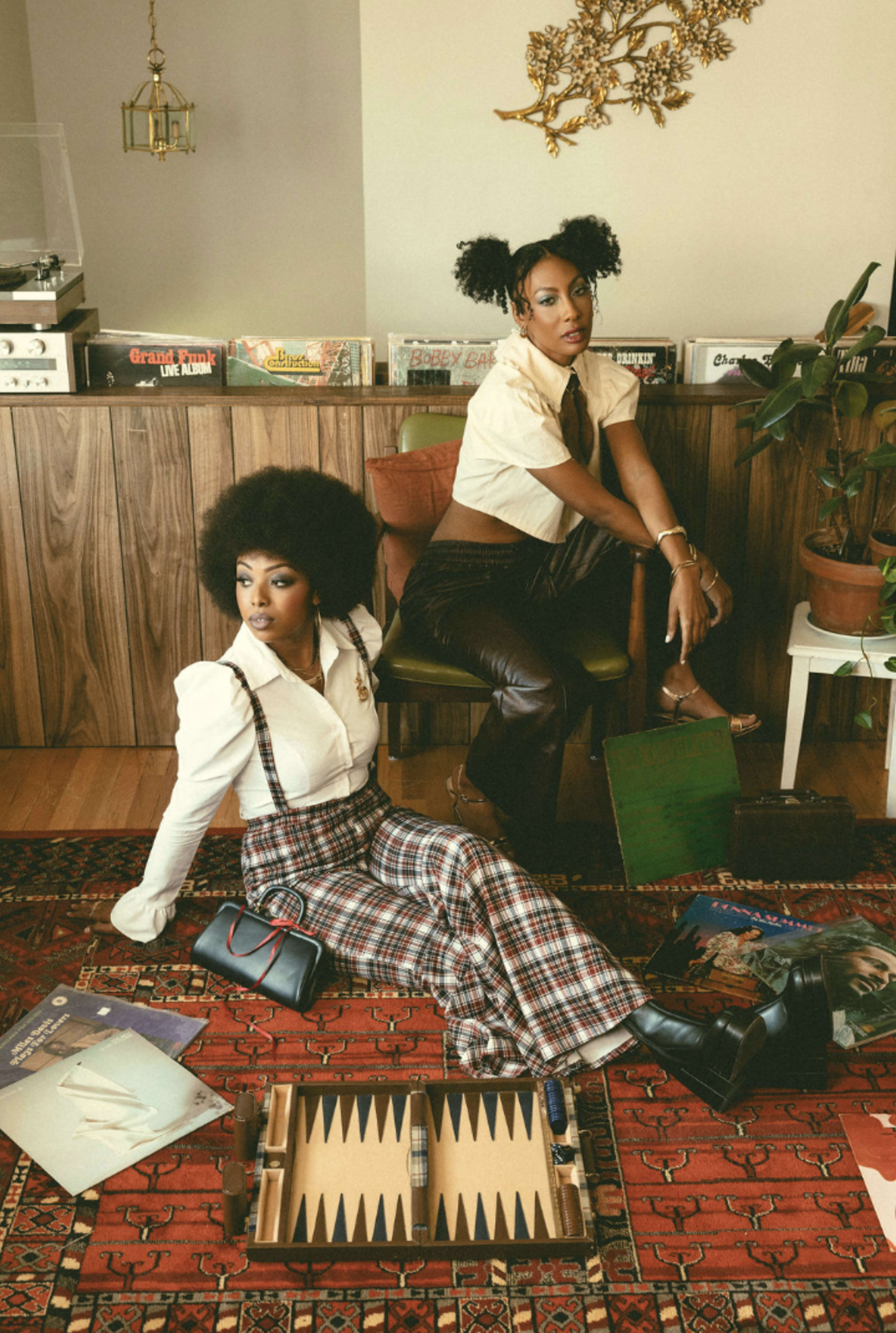 Two women with distinctive hairstyles sit on a patterned rug in a retro-styled room with vinyl records displayed in the background and a backgammon set on the floor.