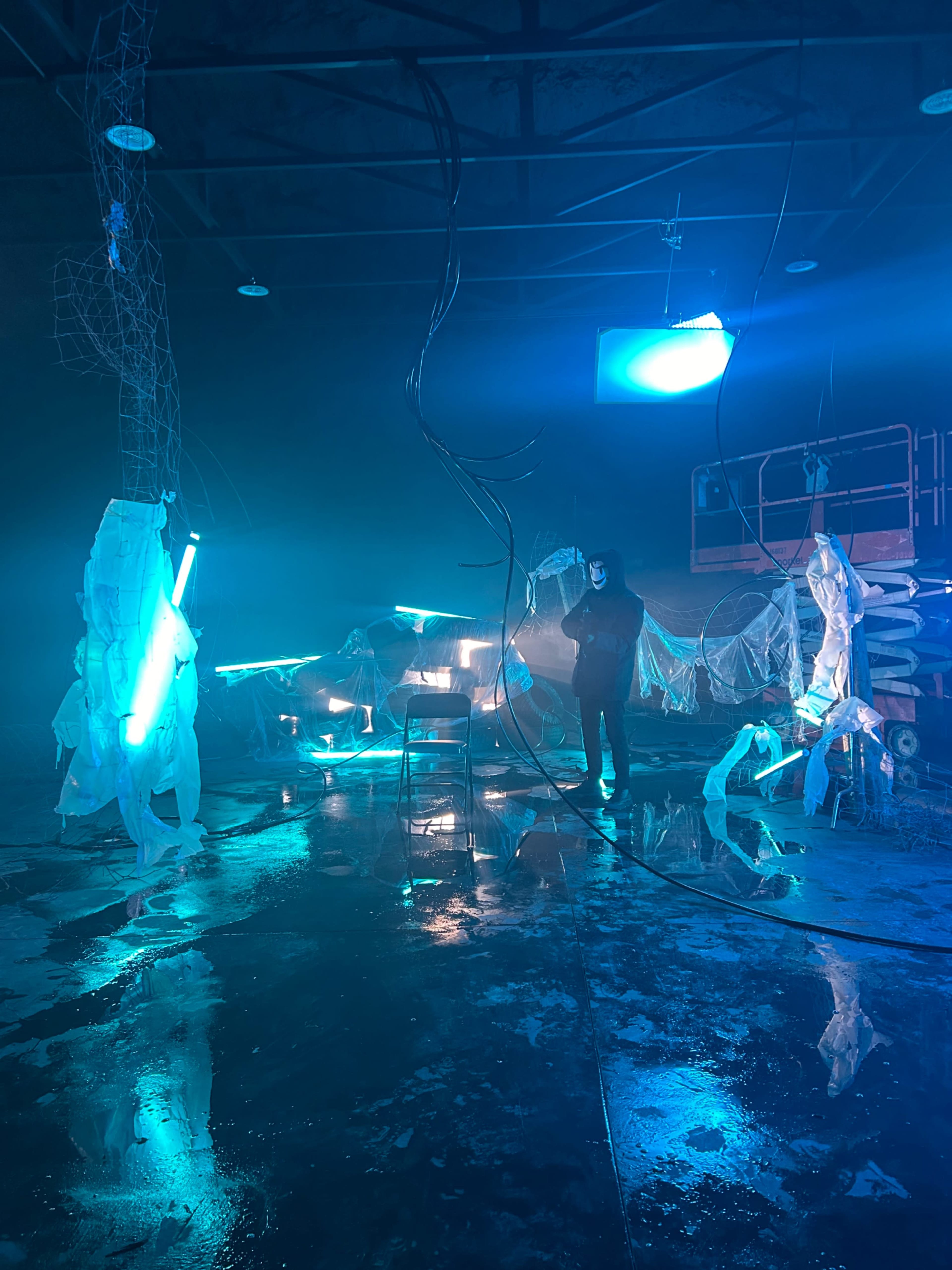 A person stands in a dimly lit industrial space surrounded by illuminated blue neon lights and reflective surfaces, with plastic sheeting and wires scattered throughout.