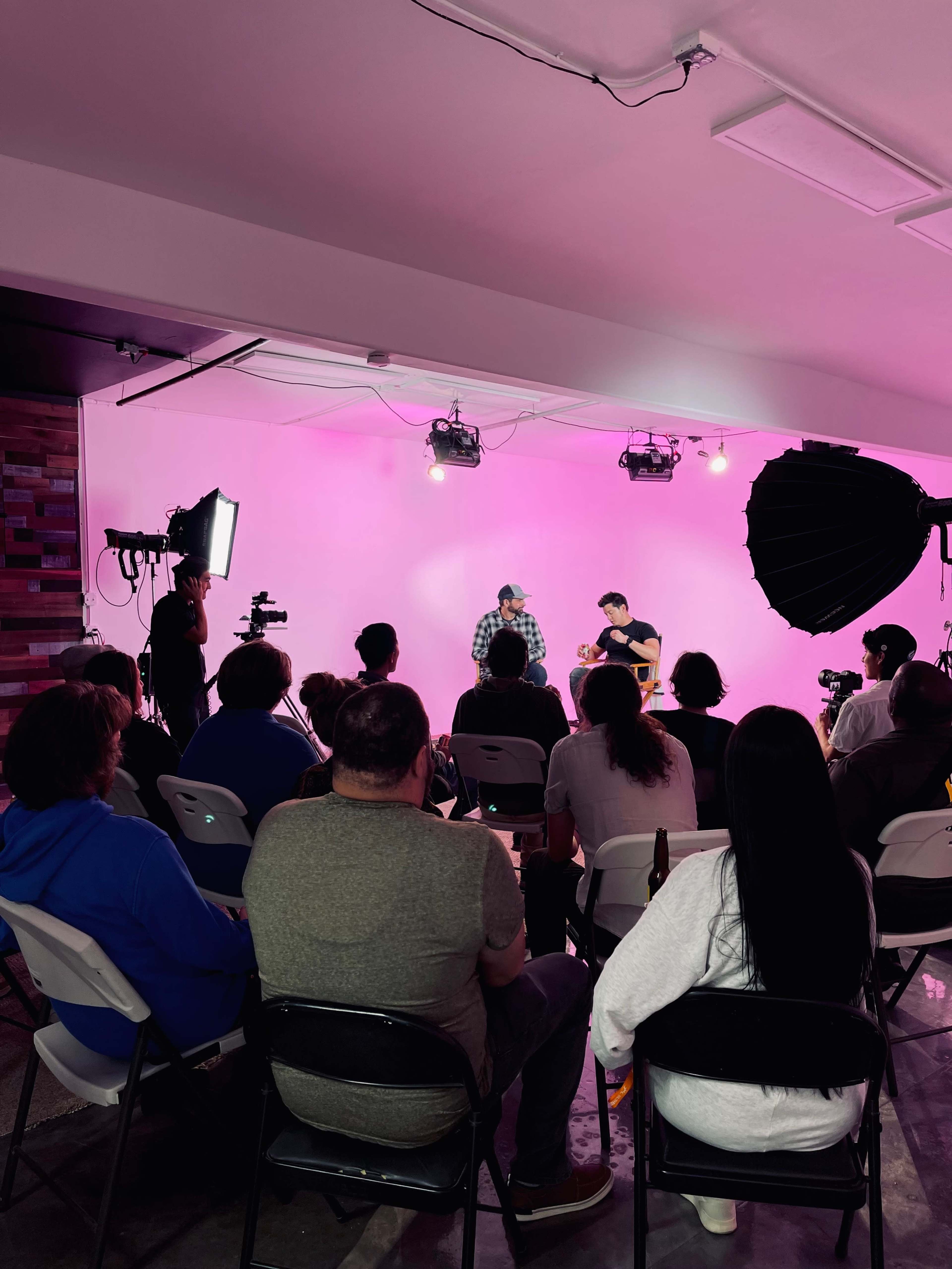 A small audience watches a live interview or performance on a stage lit with pink lighting.