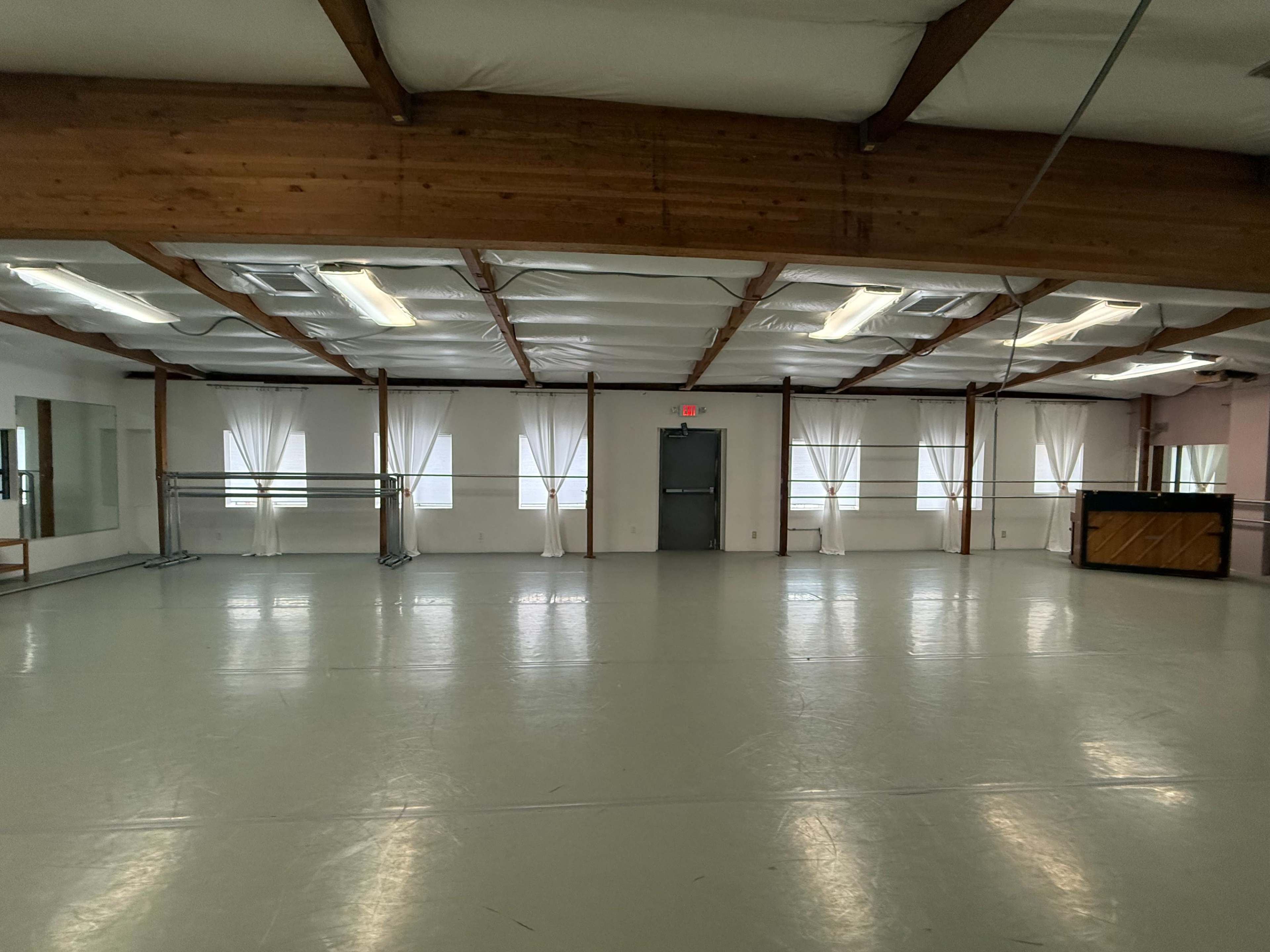 The image shows a spacious dance studio with wooden beams, large windows covered by sheer curtains, and a piano in one corner.