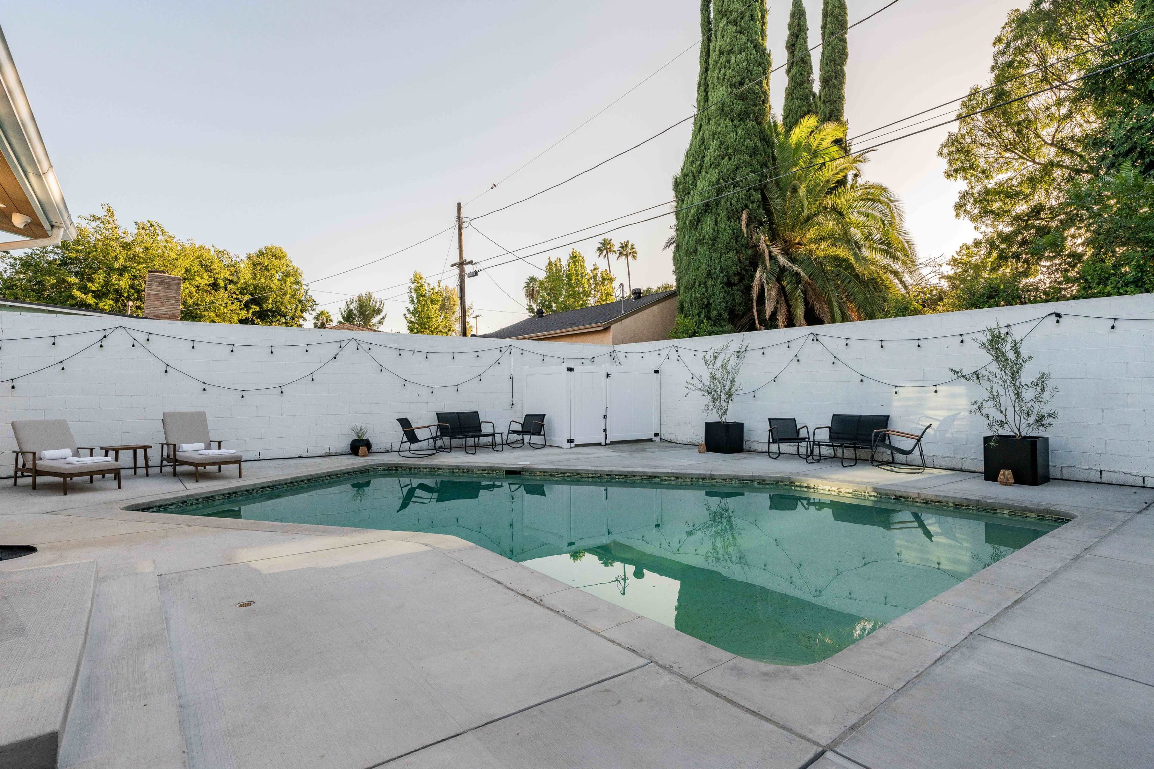 The image shows a rectangular swimming pool surrounded by a concrete patio, with several lounge chairs, trees, and string lights above.