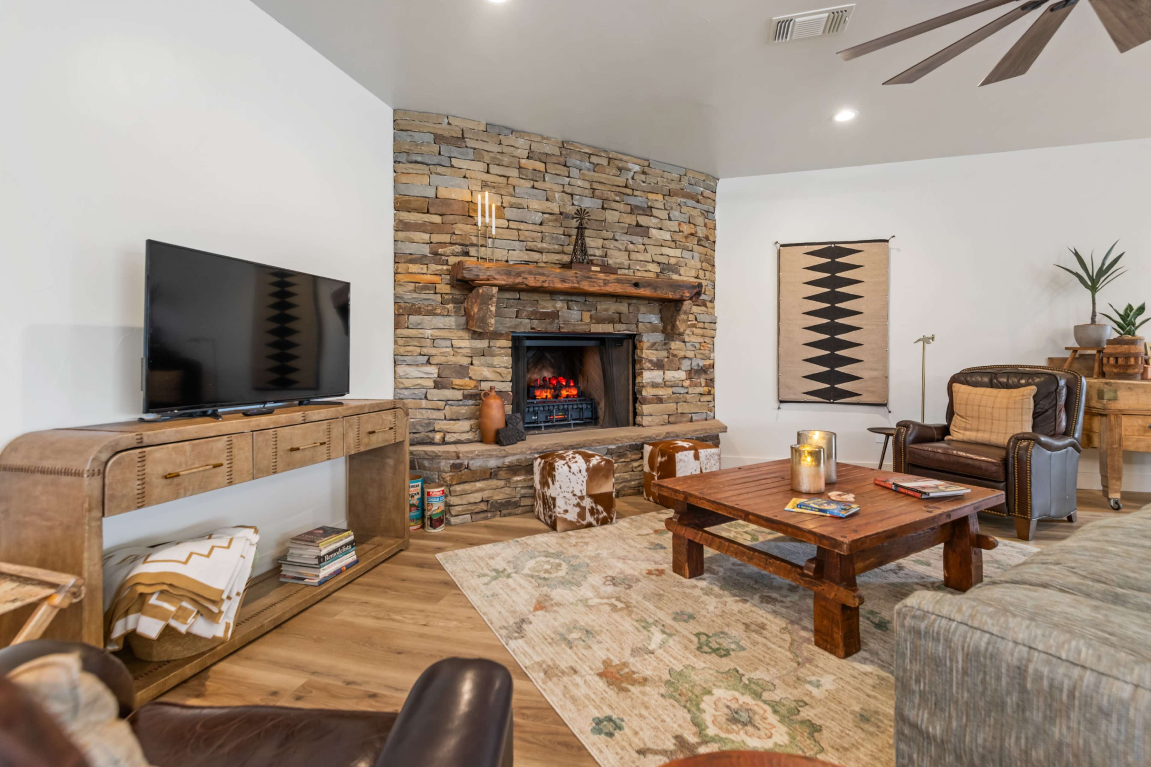 The living room features a stone fireplace with a wooden mantel, a large flat-screen TV, and a mix of comfortable seating around a wooden coffee table.