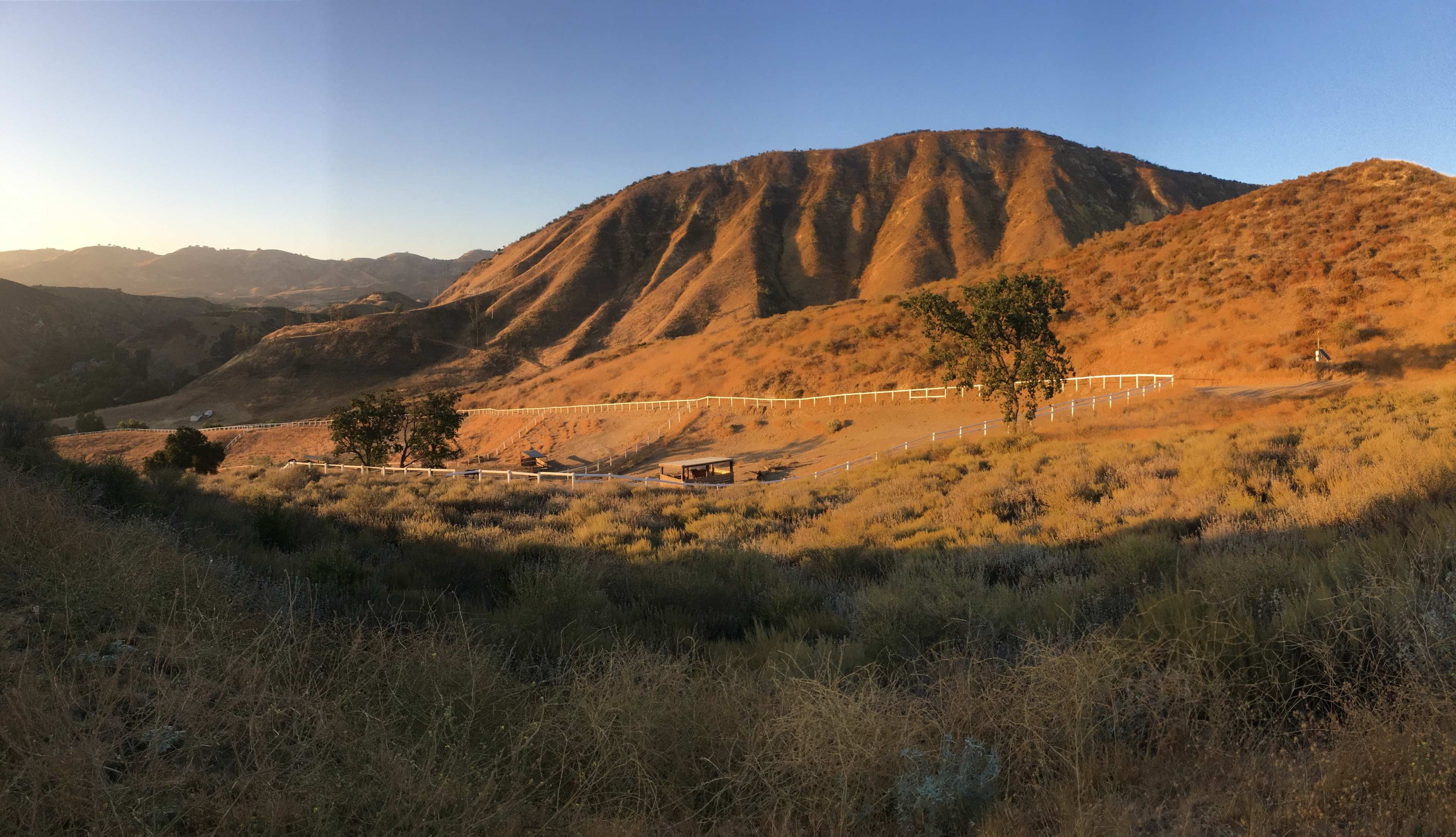 The image shows a hilly landscape with golden sunlight illuminating the terrain and a white fence outlining a field in the foreground.