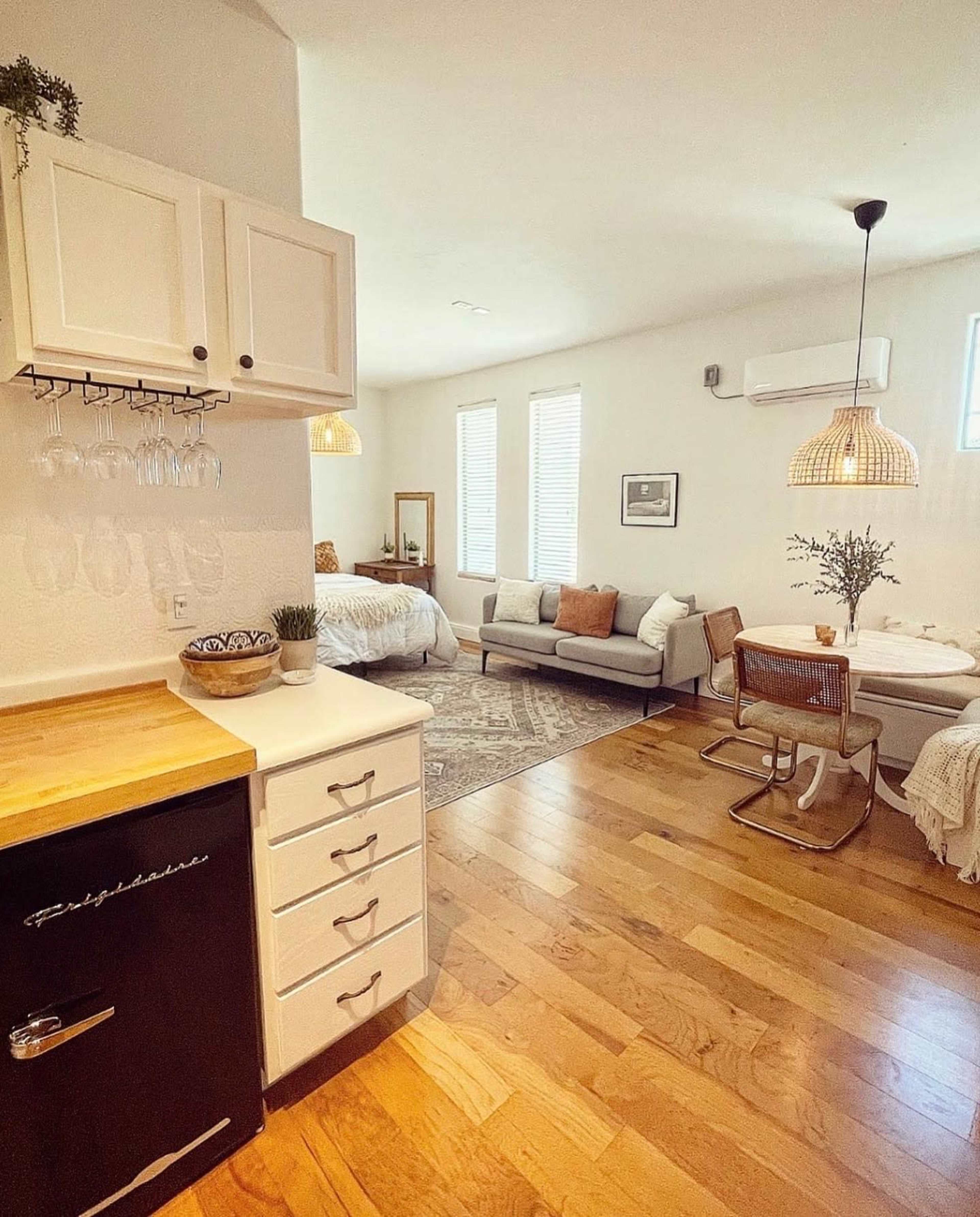 A small, open-concept living space features a kitchenette, seating area, and a bedroom with natural light streaming through windows.