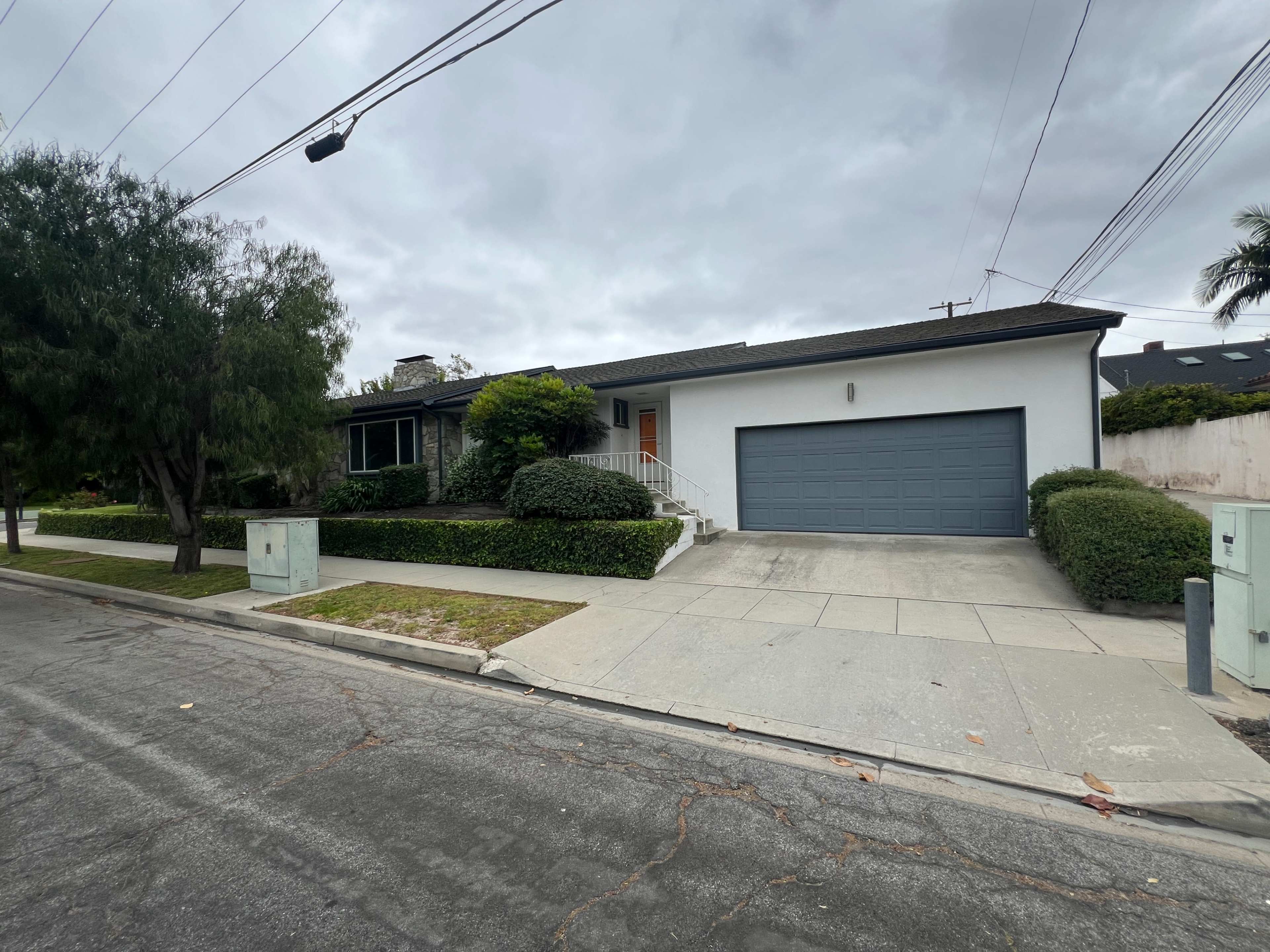 The image shows a suburban single-story house with a grey garage door, a walkway leading to the front door, and landscaped greenery in the yard.