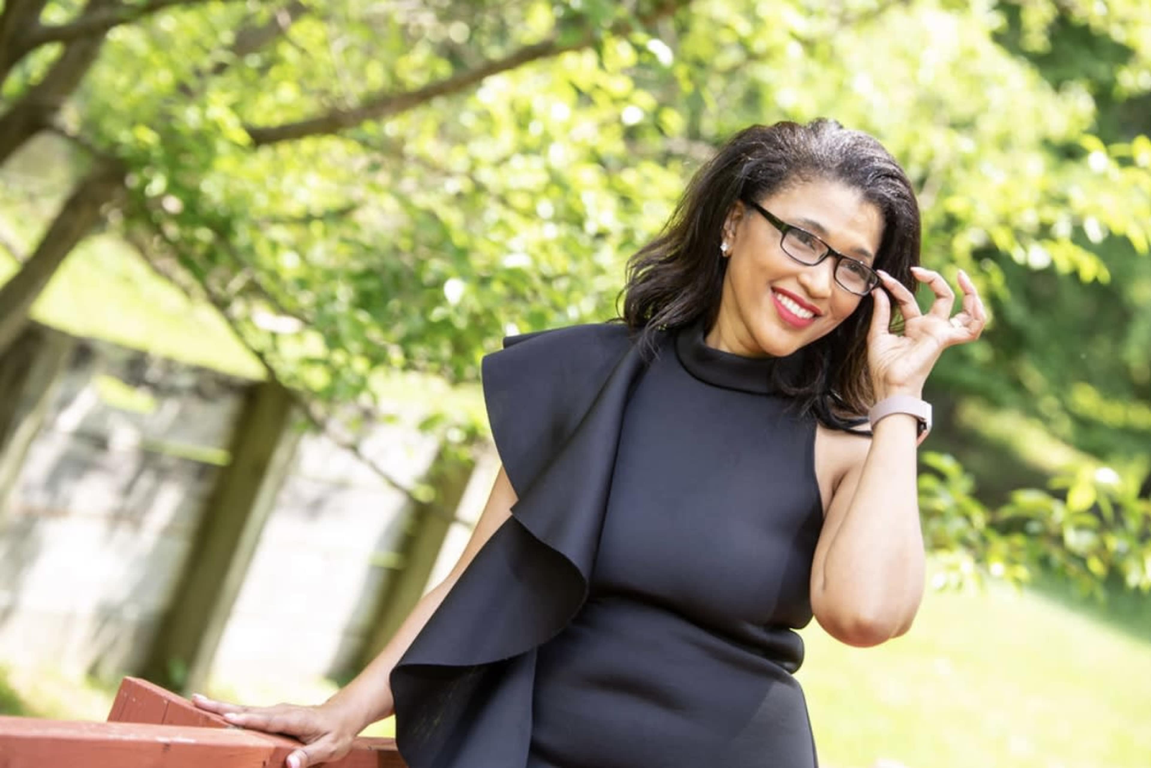 A woman in a black dress stands outdoors, smiling while adjusting her glasses.