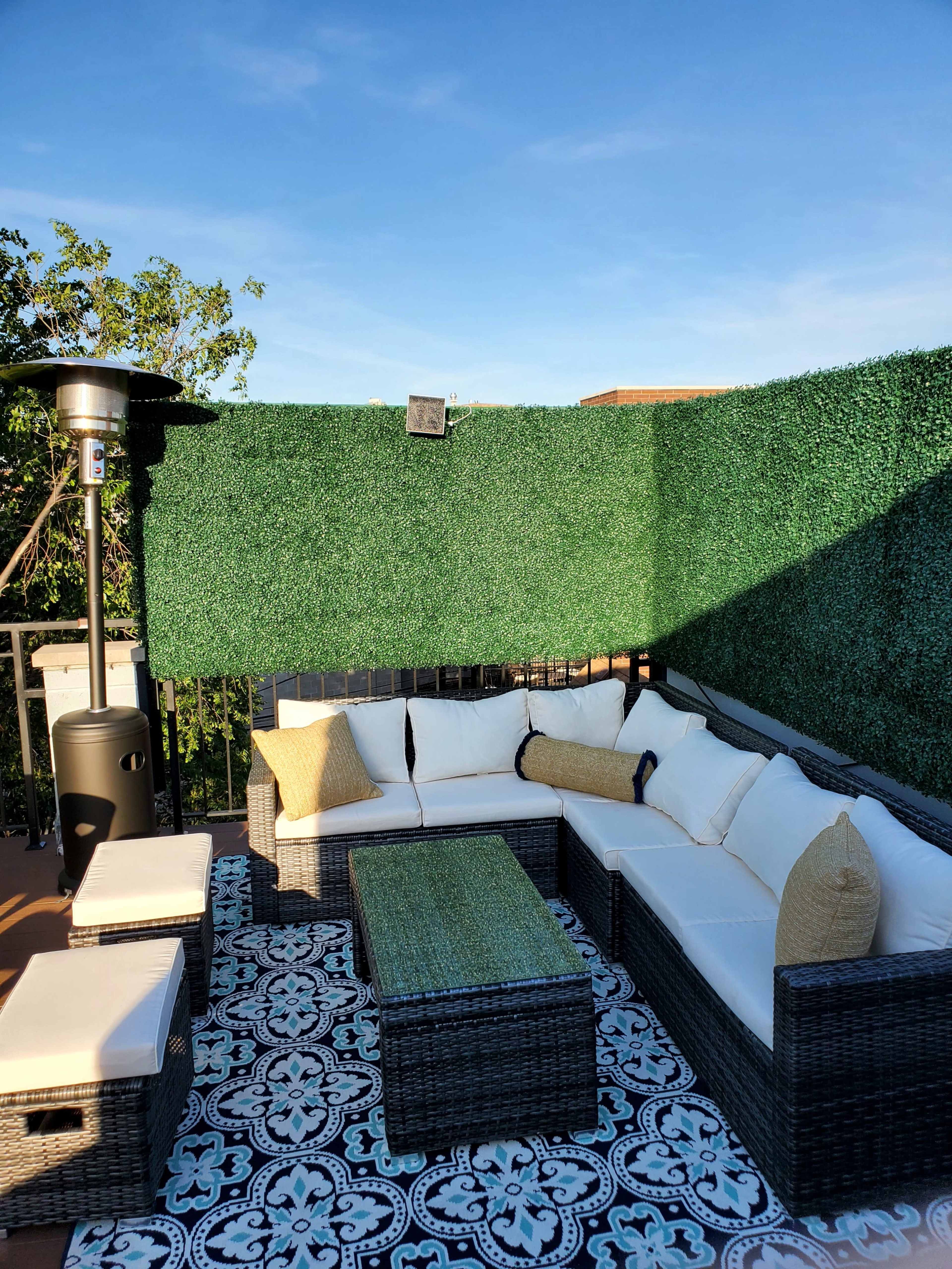 A rooftop patio features a sectional sofa with white cushions, a green area rug, and a heater surrounded by tall artificial hedges.