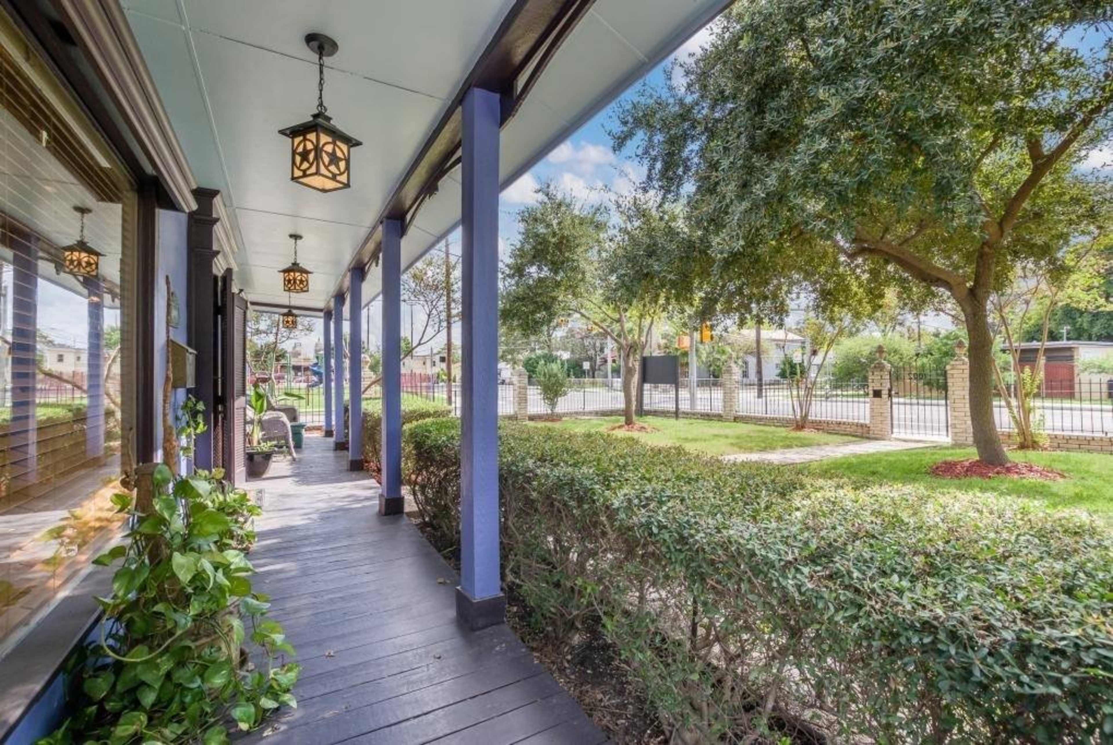 A porch area with hanging lanterns, bordered by manicured bushes, leading to a view of a street beyond.