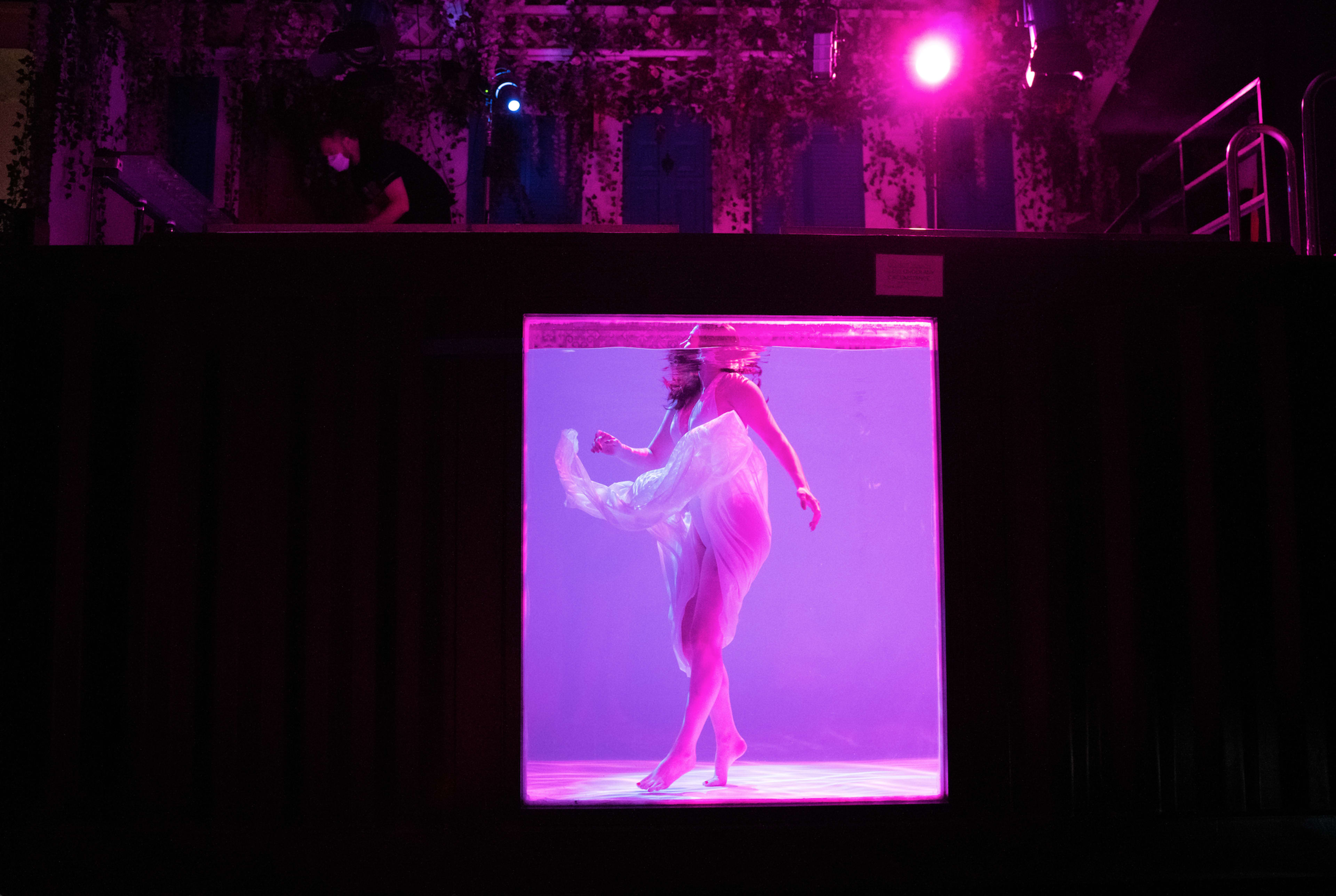 A performer dances in a submerged glass enclosure while a person operates equipment above in a dimly lit venue.