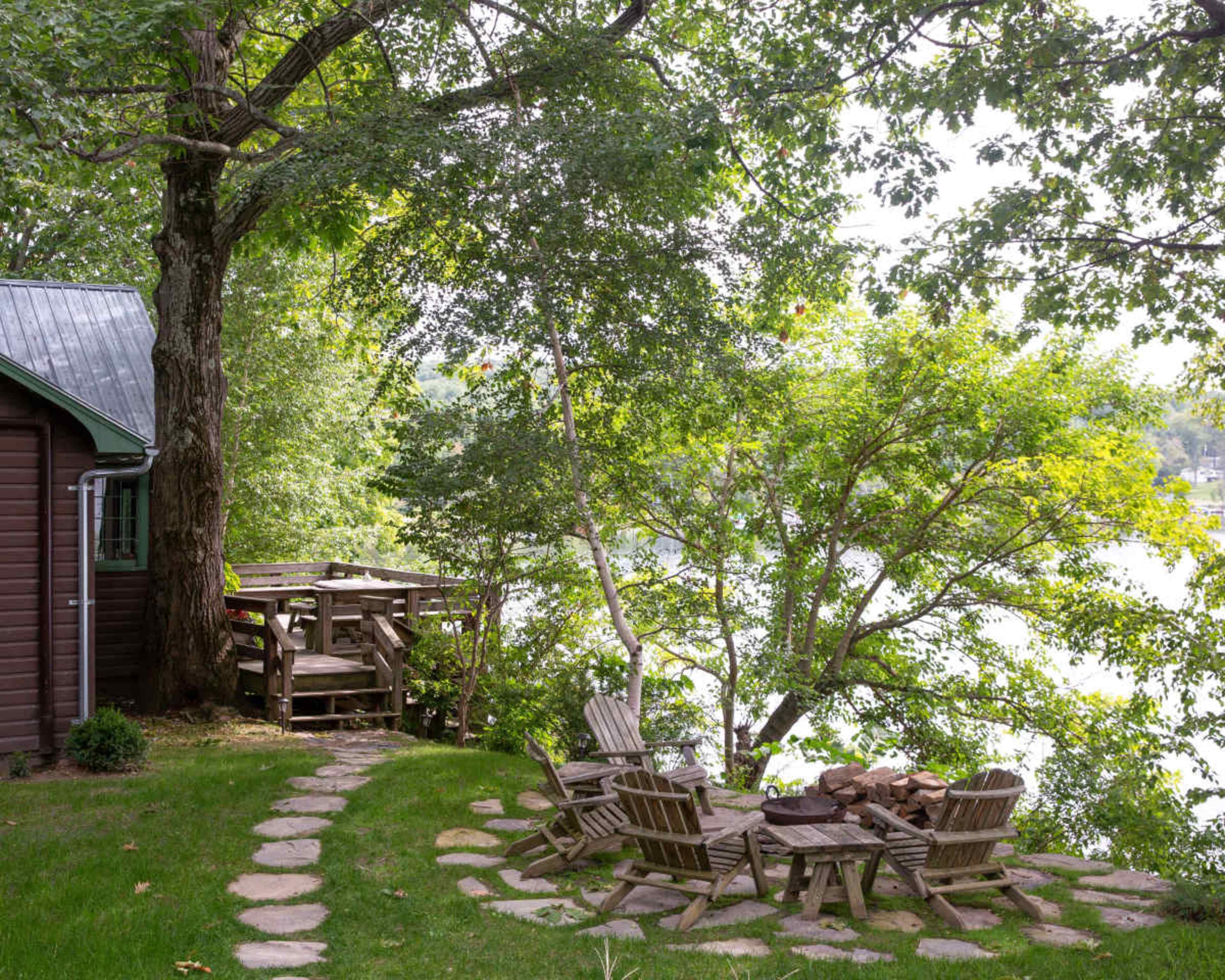 A circular stone patio with wooden chairs surrounds a fire pit near a lake, with a tree providing shade and a wooden deck visible nearby.