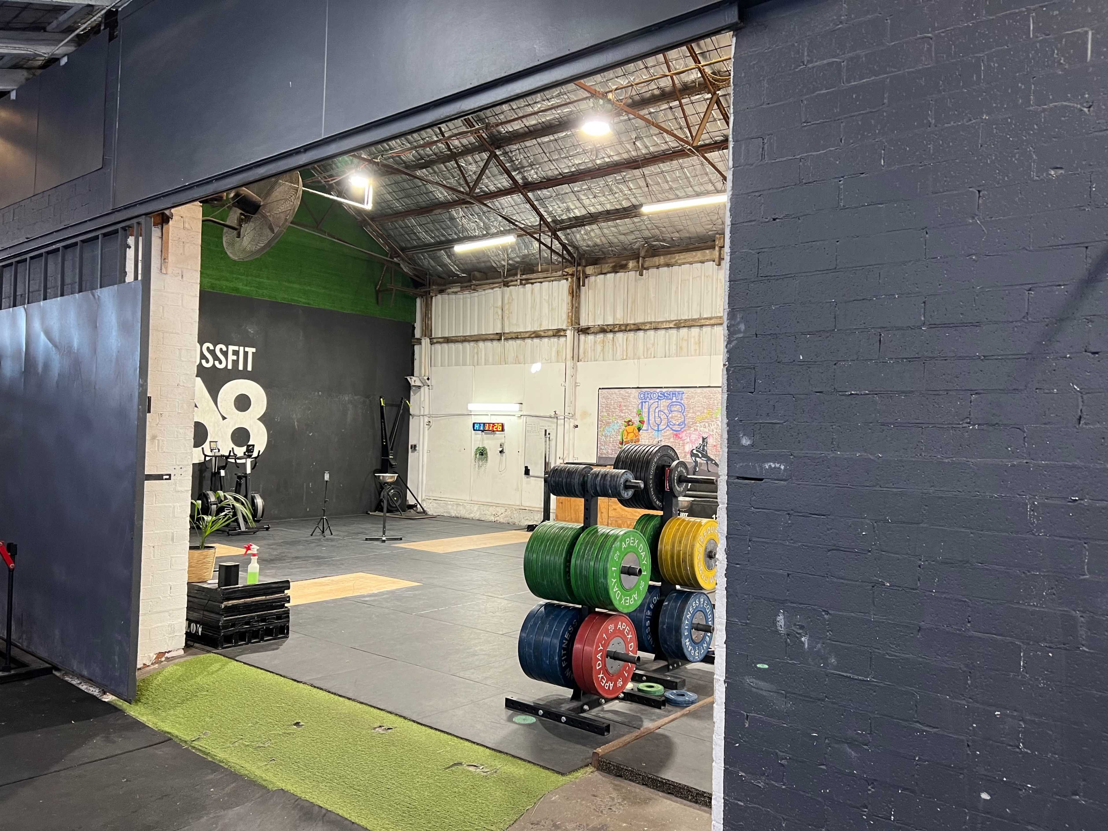 A view of a CrossFit gym with weightlifting equipment and a training area visible through an open doorway.