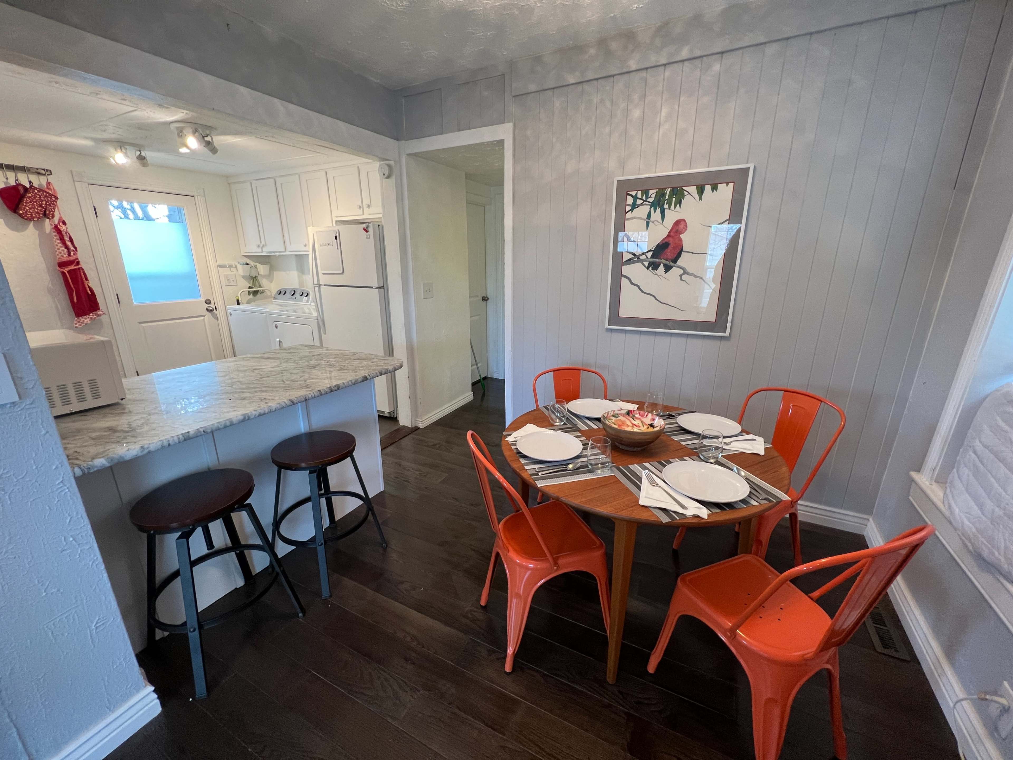 A small dining area features a round wooden table set for four, surrounded by orange metal chairs, with a view of a kitchen area in the background.